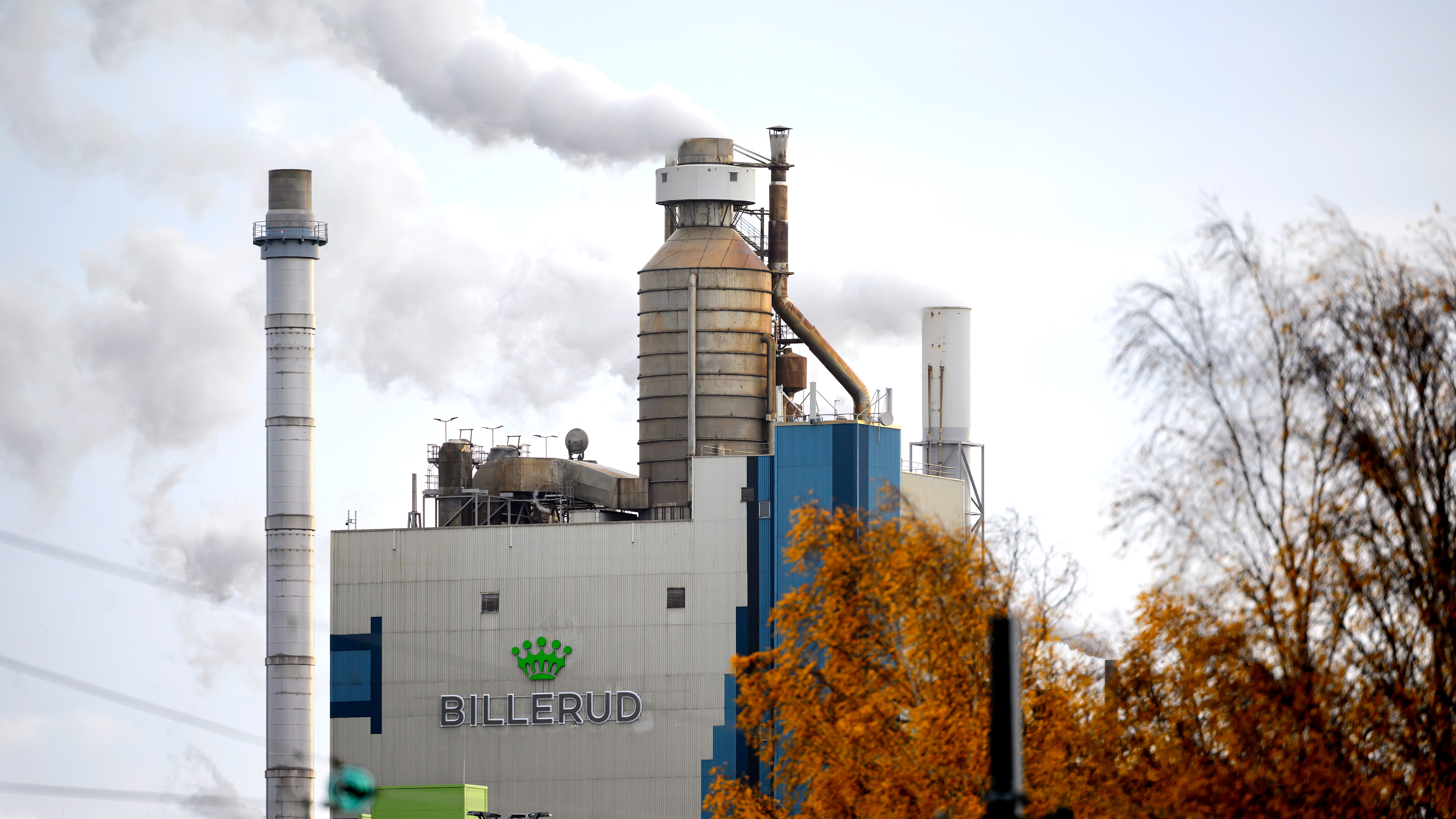 Industribyggnad med texten "Billerud" som släpper ut vit rök från skorstenarna, träd syns i förgrunden.