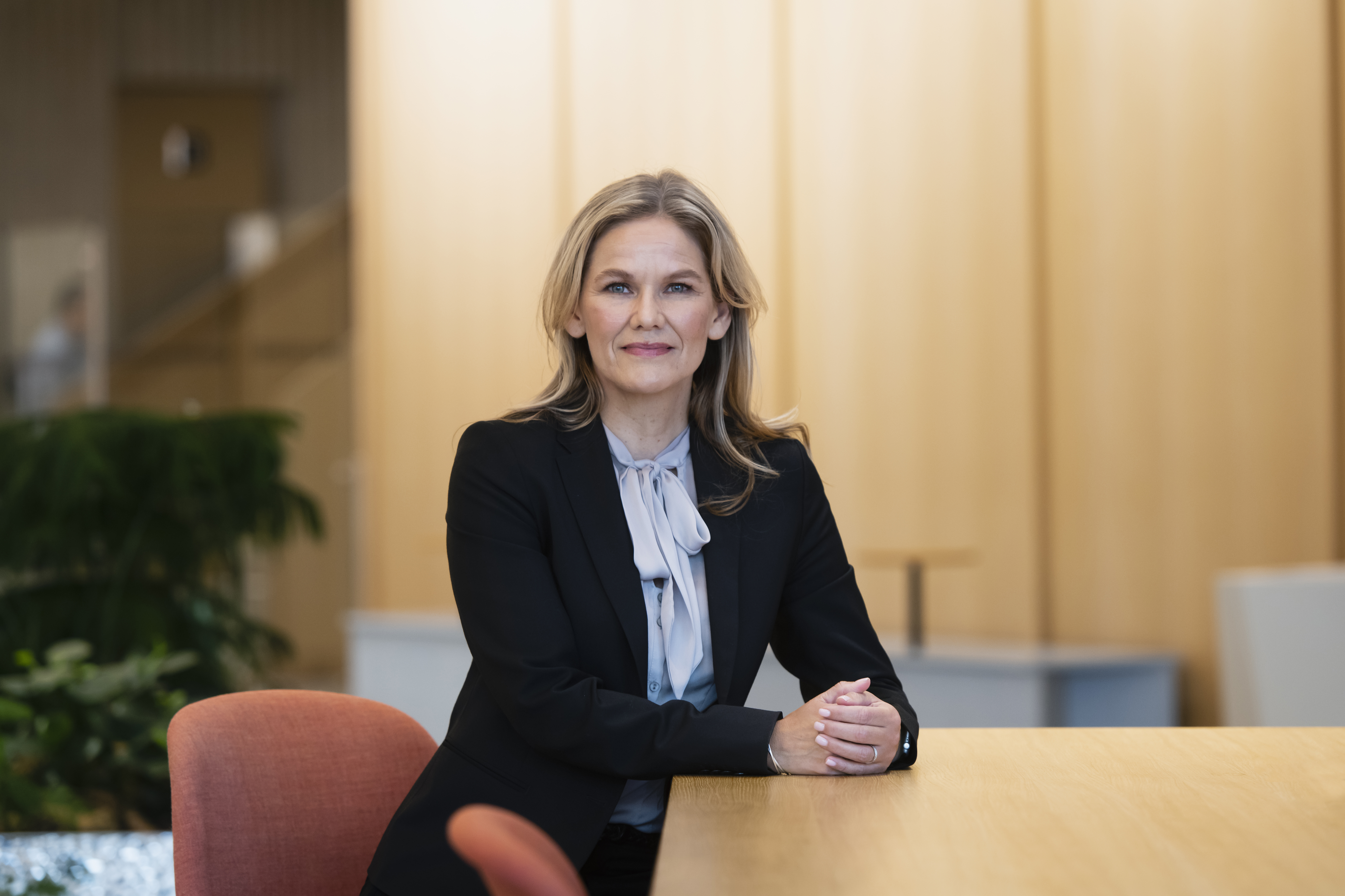 En kvinna i svart kavaj sitter vid ett träbord på ett kontor med ljusa väggar och växter i bakgrunden.