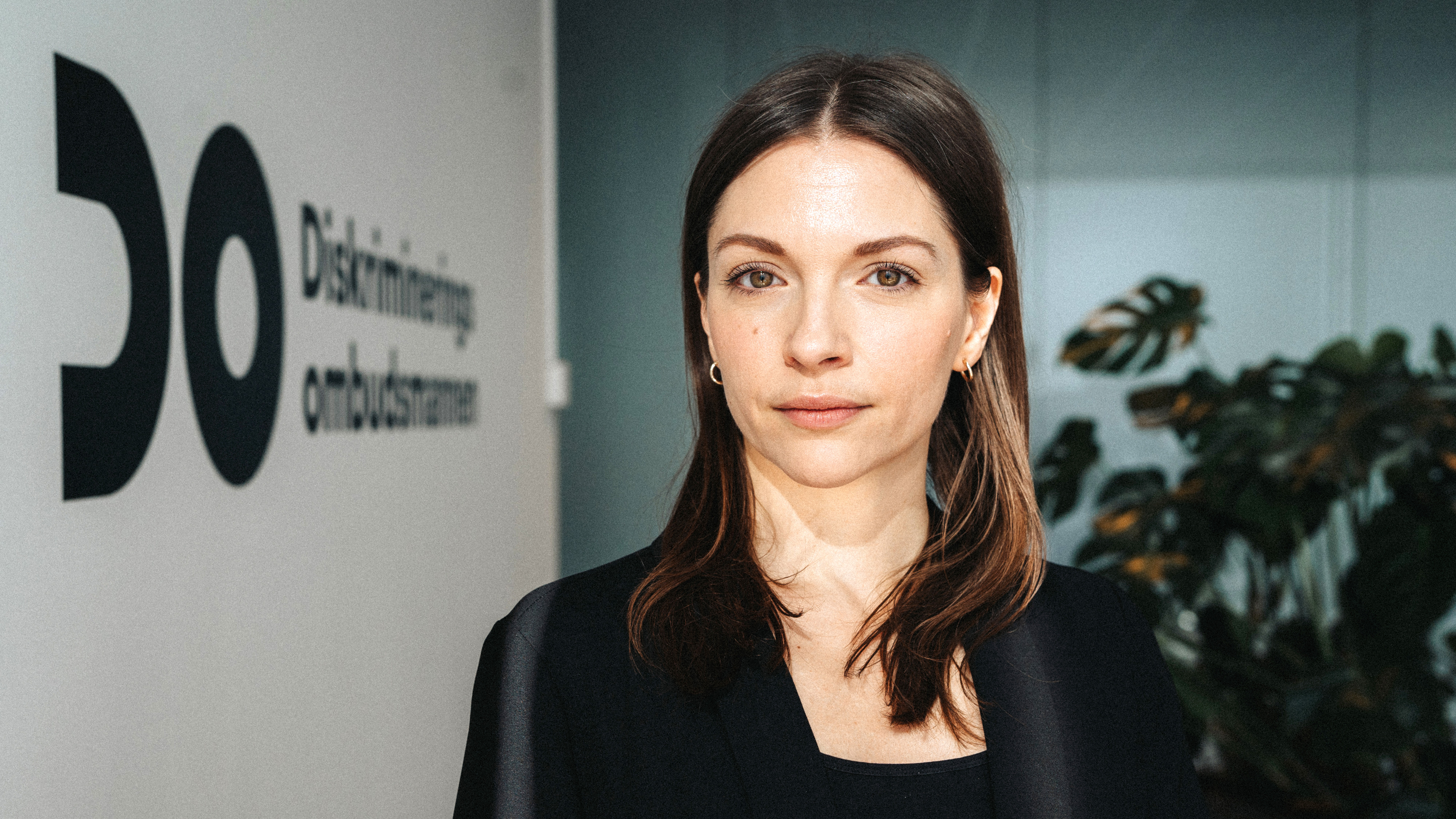 En kvinna med rakt brunt hår och svart kavaj står inomhus vid en vägg med texten "Diskriminerings ombudsmannen" och en stor växt.