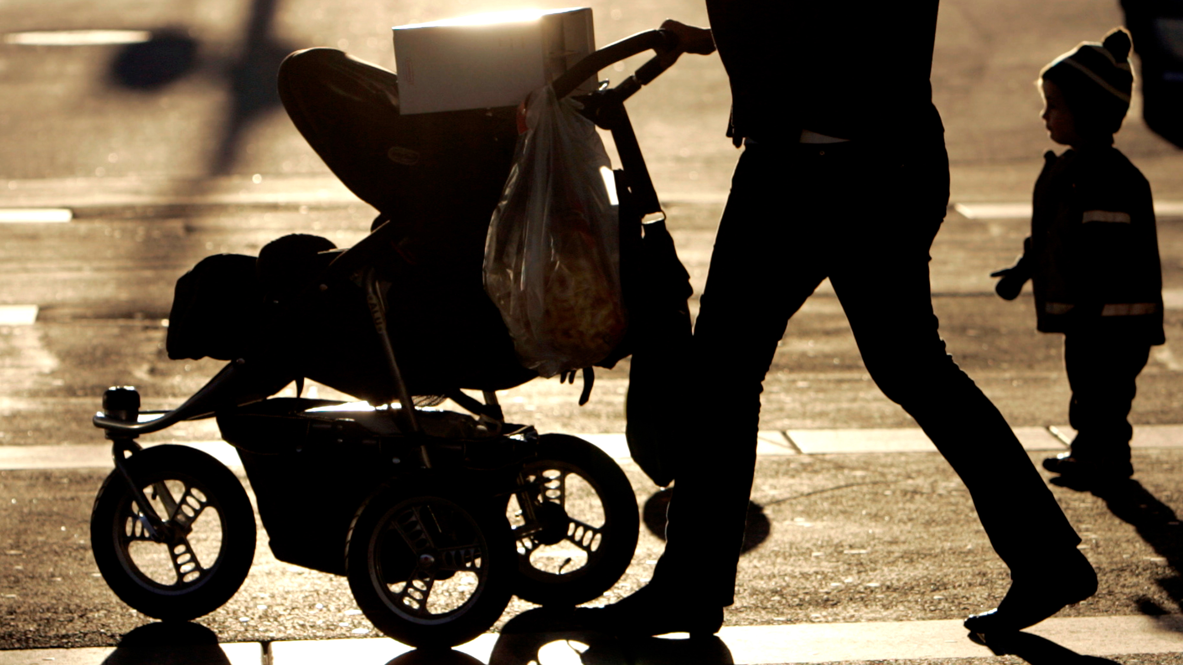 En person skjuter en barnvagn med en plastpåse hängande från den medan ett litet barn går bredvid på en gata.