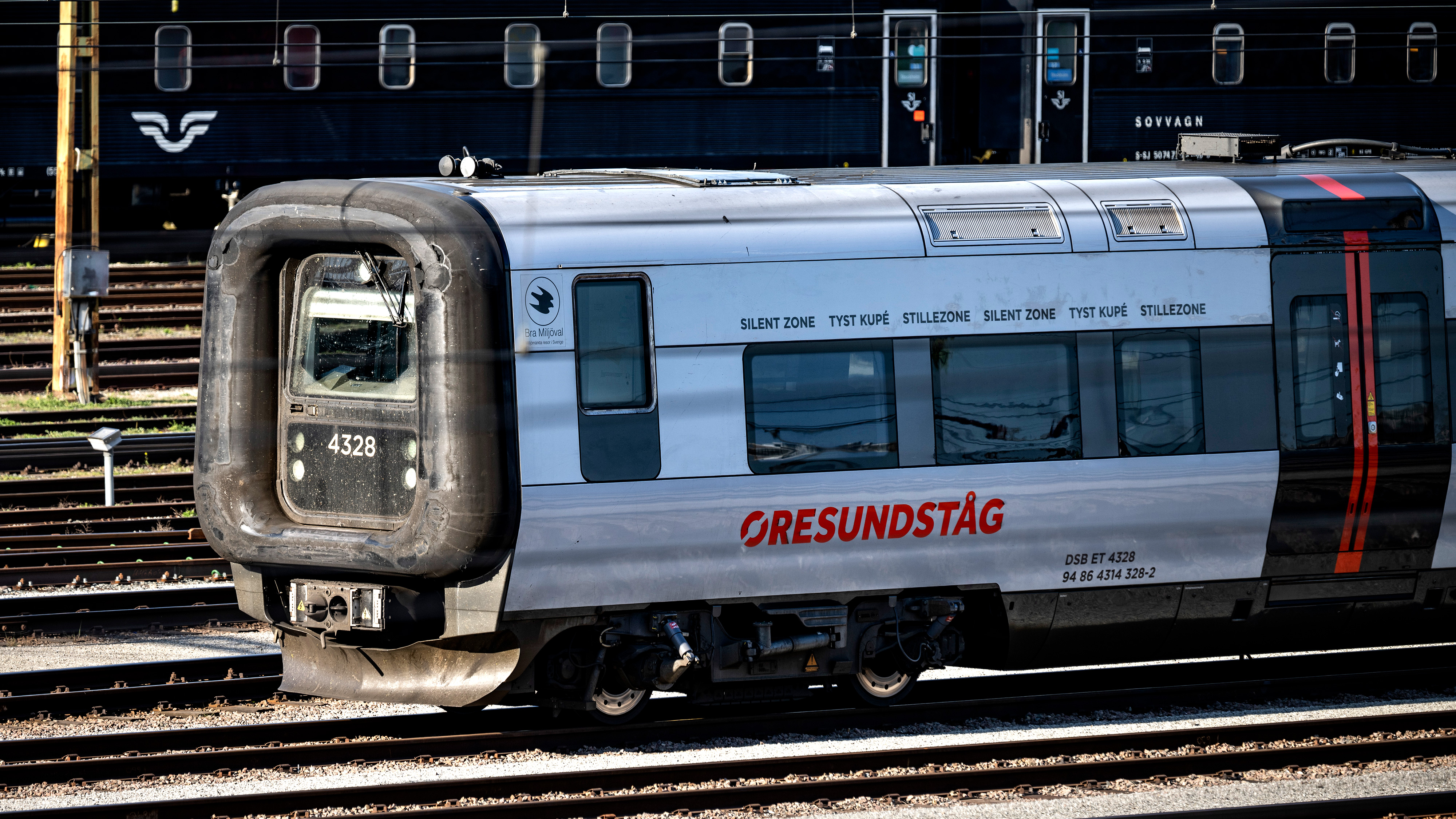 Ett Öresundståg med nummer 4328 står på järnvägsspår, och ett svart tåg syns i bakgrunden.