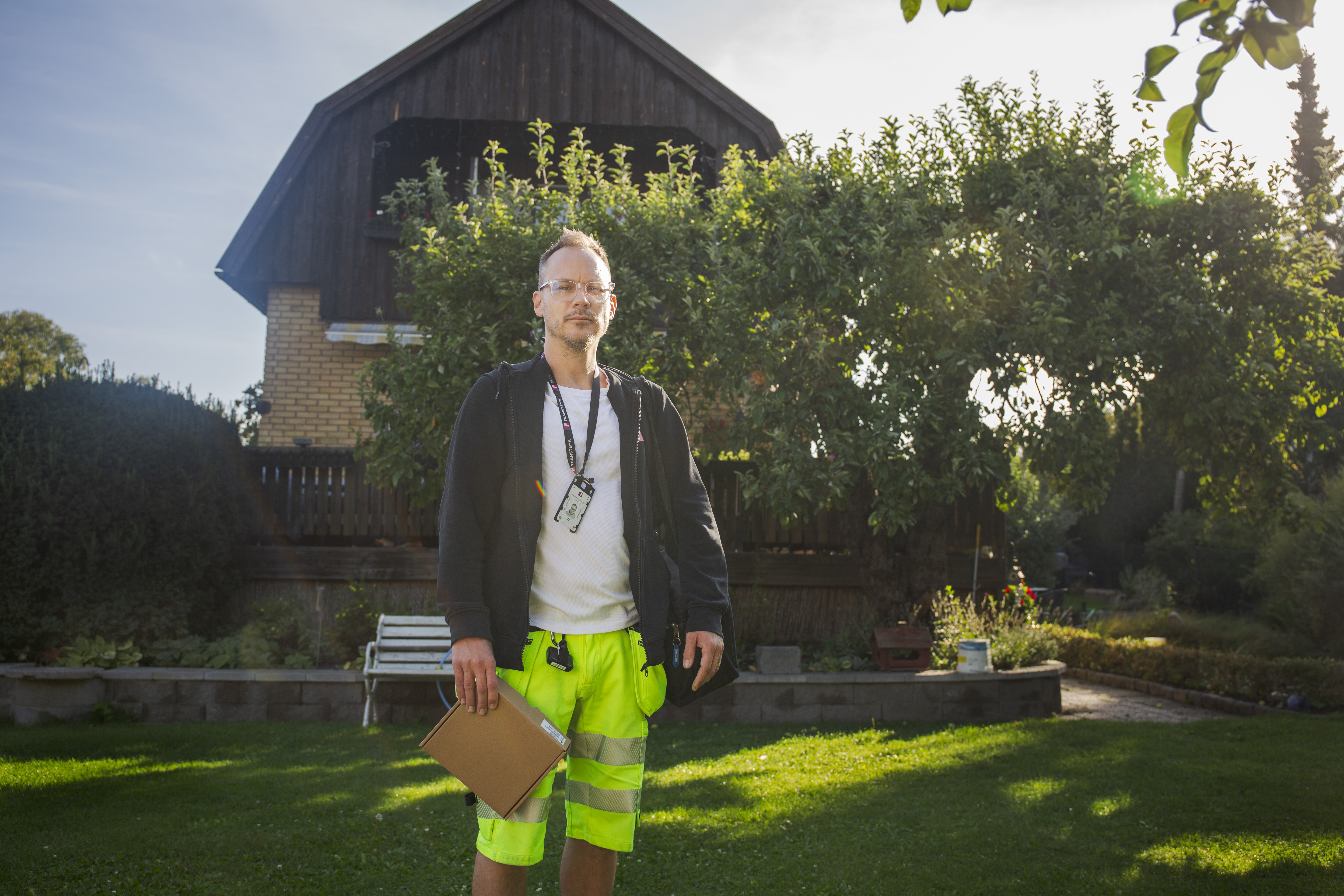 En man med svart jacka och neongula arbetsshorts står på en gräsmatta med ett block, ett hus och träd syns i bakgrunden.