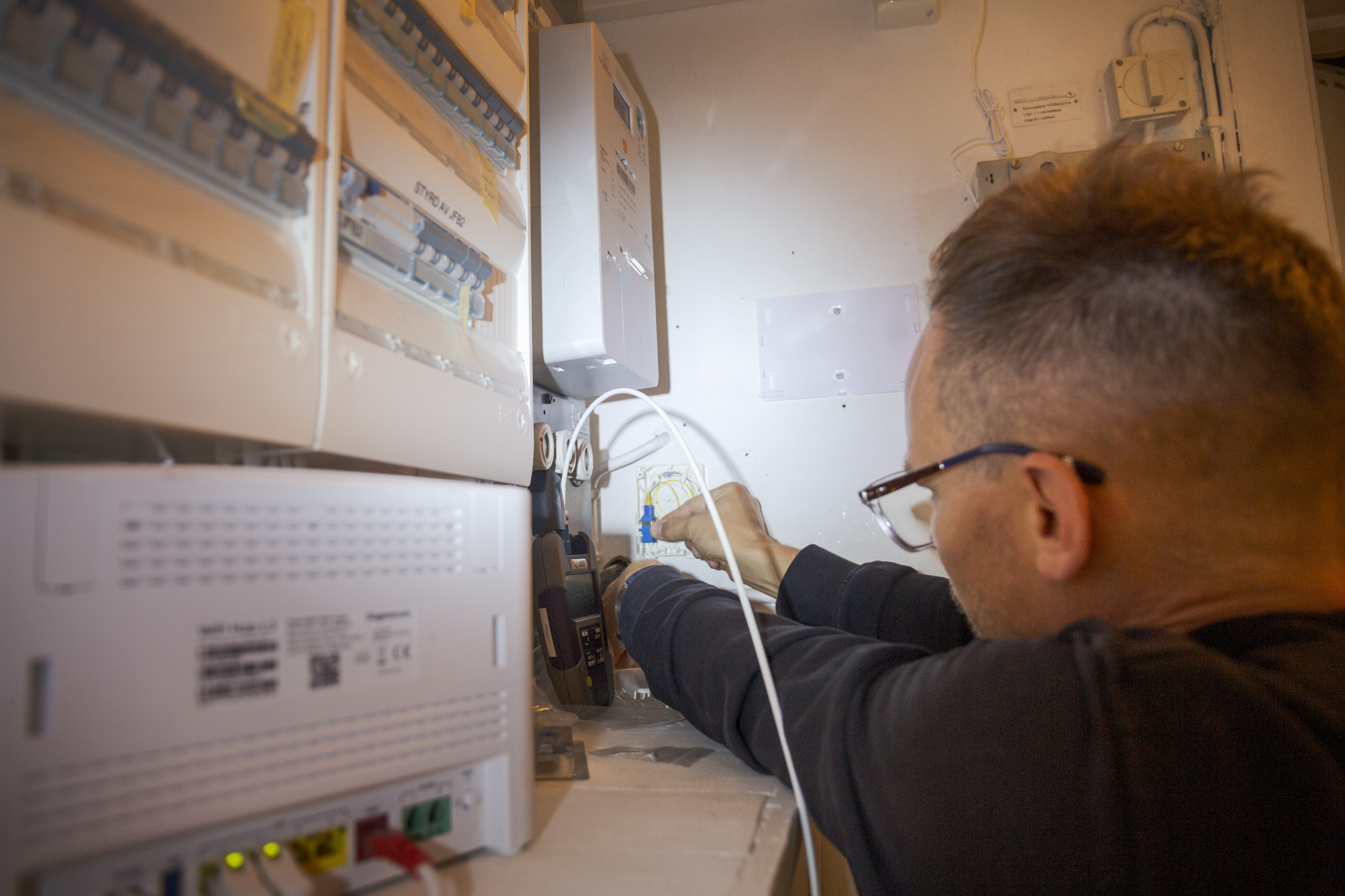 En person med glasögon installerar eller reparerar elektriska komponenter i en kopplingsdosa bland sladdar och utrustning.