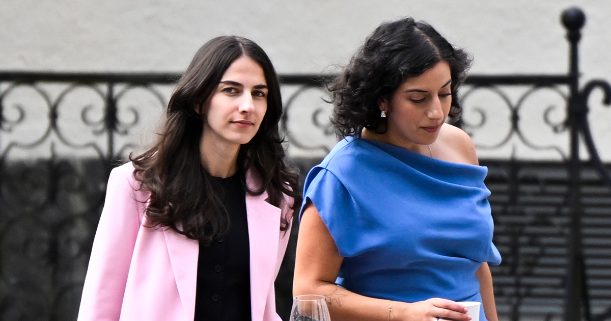 Två kvinnor går utomhus; en har ljusrosa kostym och svart topp, den andra har blå axelklänning och håller ett glas.