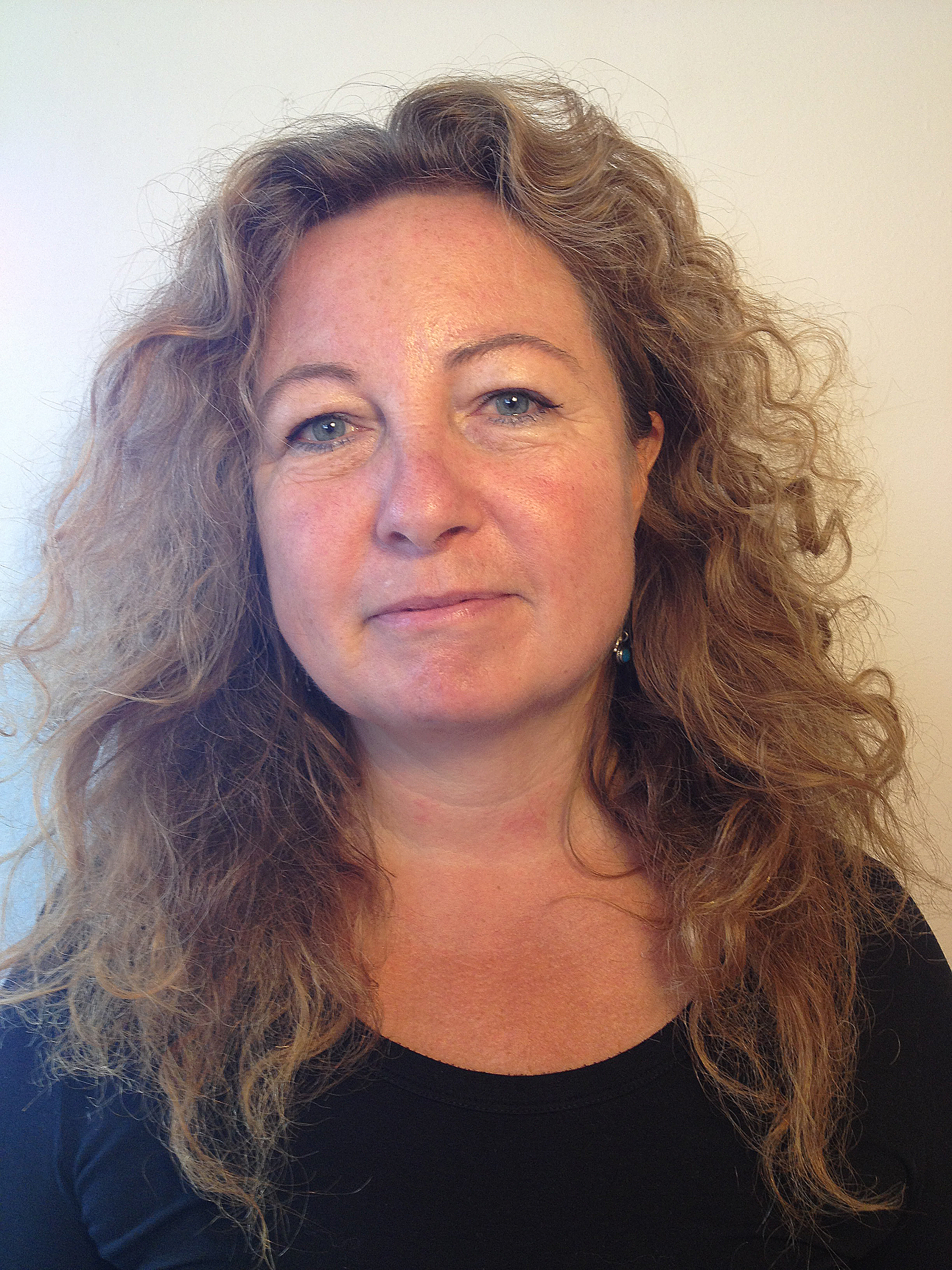 Linda Kjaer Minke med blont, vågigt hår och blå ögon har en svart topp och står framför en ljus bakgrund.