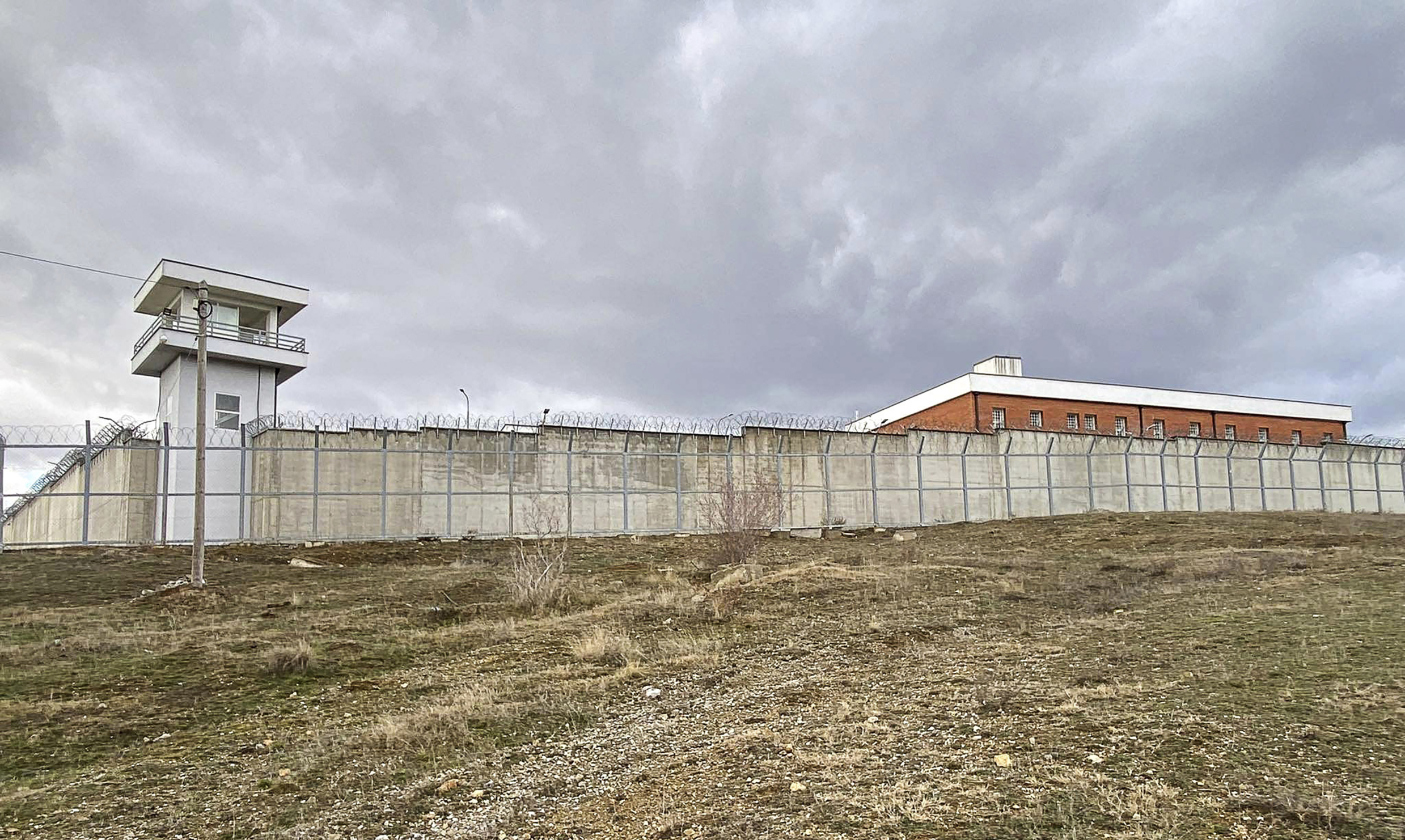 Ett fängelseområde med stängsel och taggtråd, ett vakttorn, betongmurar och en byggnad i rött tegel under molnig himmel.