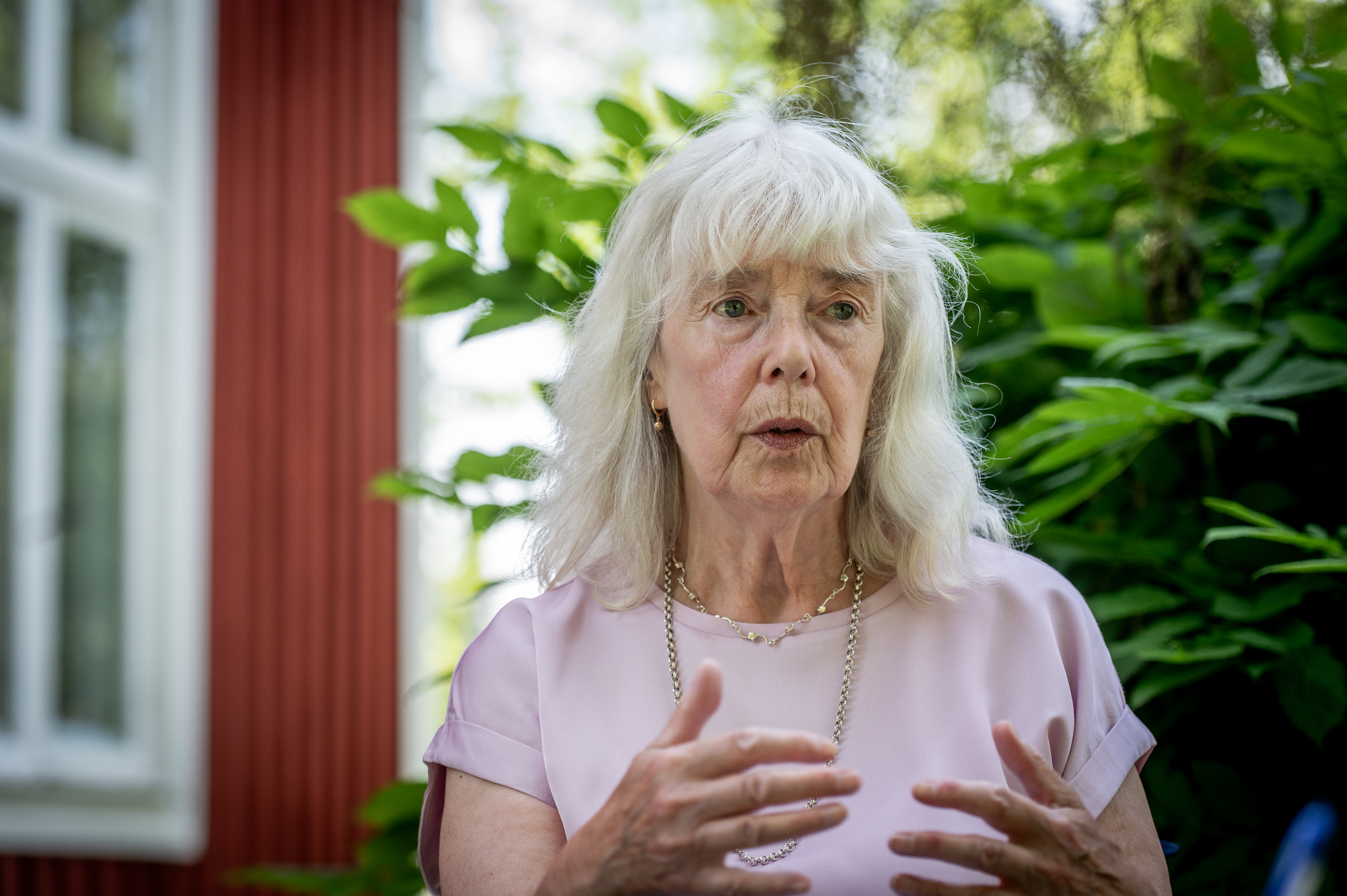Annette Thörnquist med långt grått hår och en ljusrosa blus gestikulerar med händerna utomhus nära gröna växter och en röd byggnad.