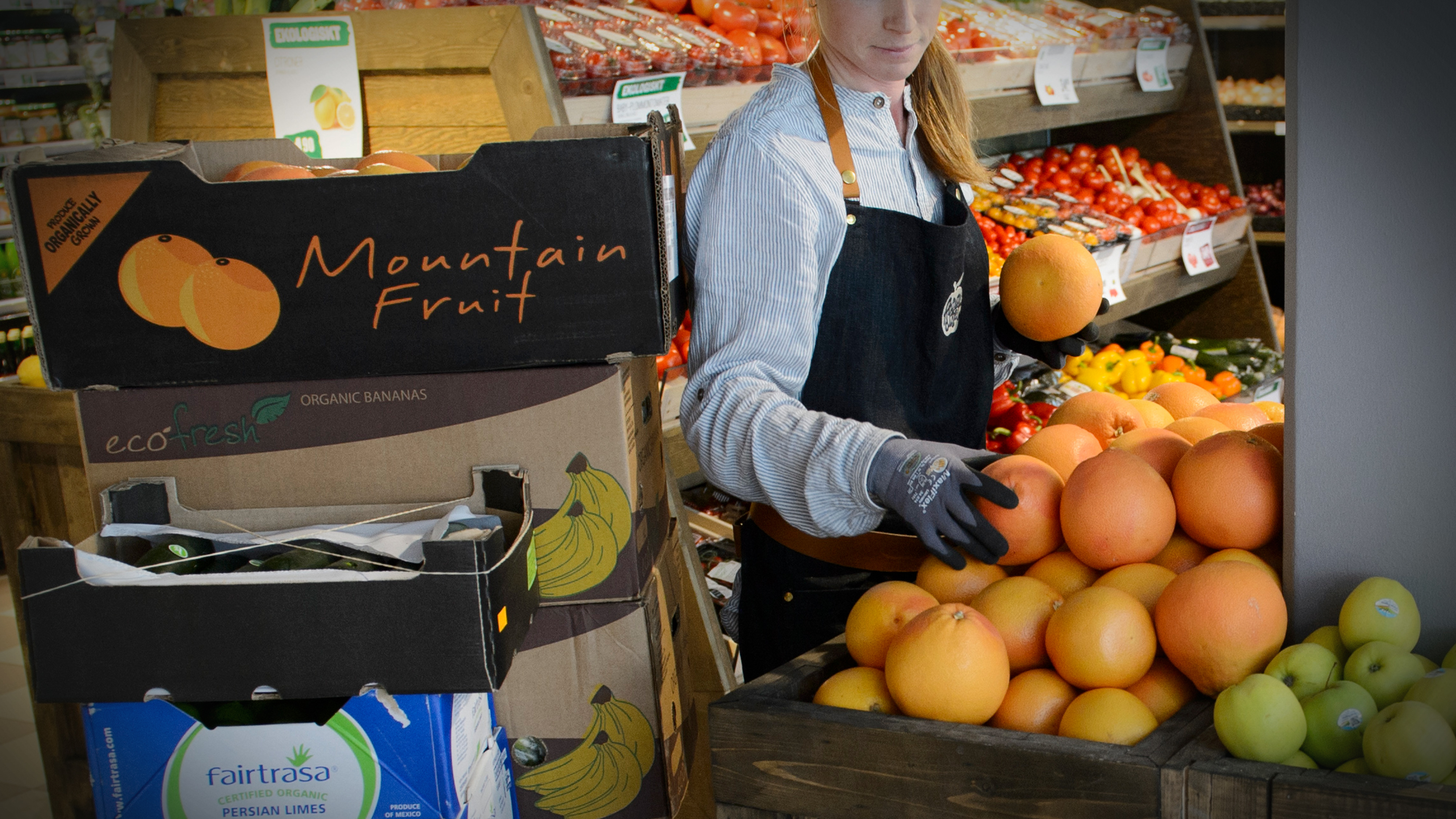 En butiksanställd med handskar ordnar grapefrukt i fruktavdelningen, med lådor bananer och lime bredvid.