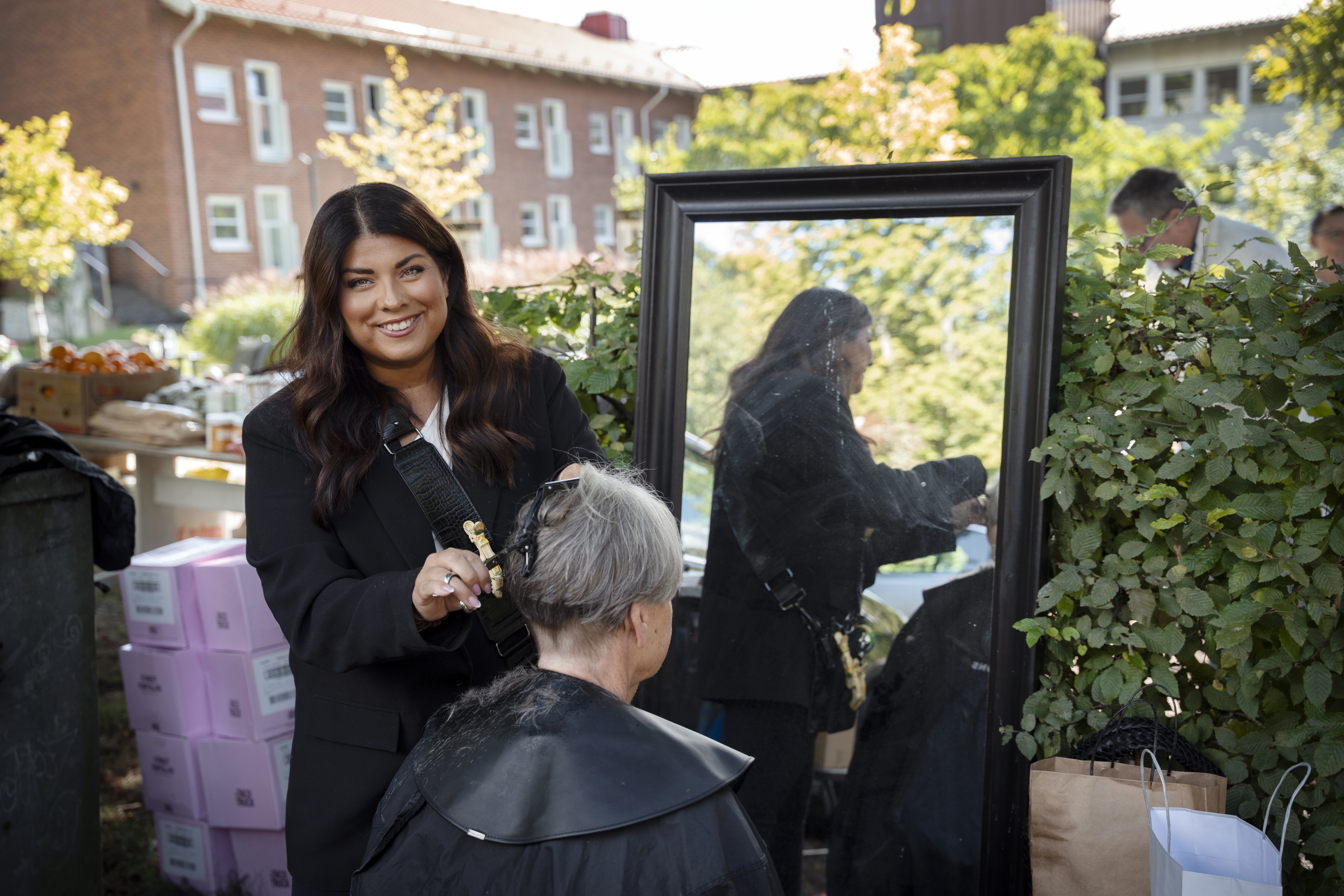 En kvinna klipper en annan person hår utomhus bredvid en spegel, omgiven av växter och lådor, med byggnader i bakgrunden.