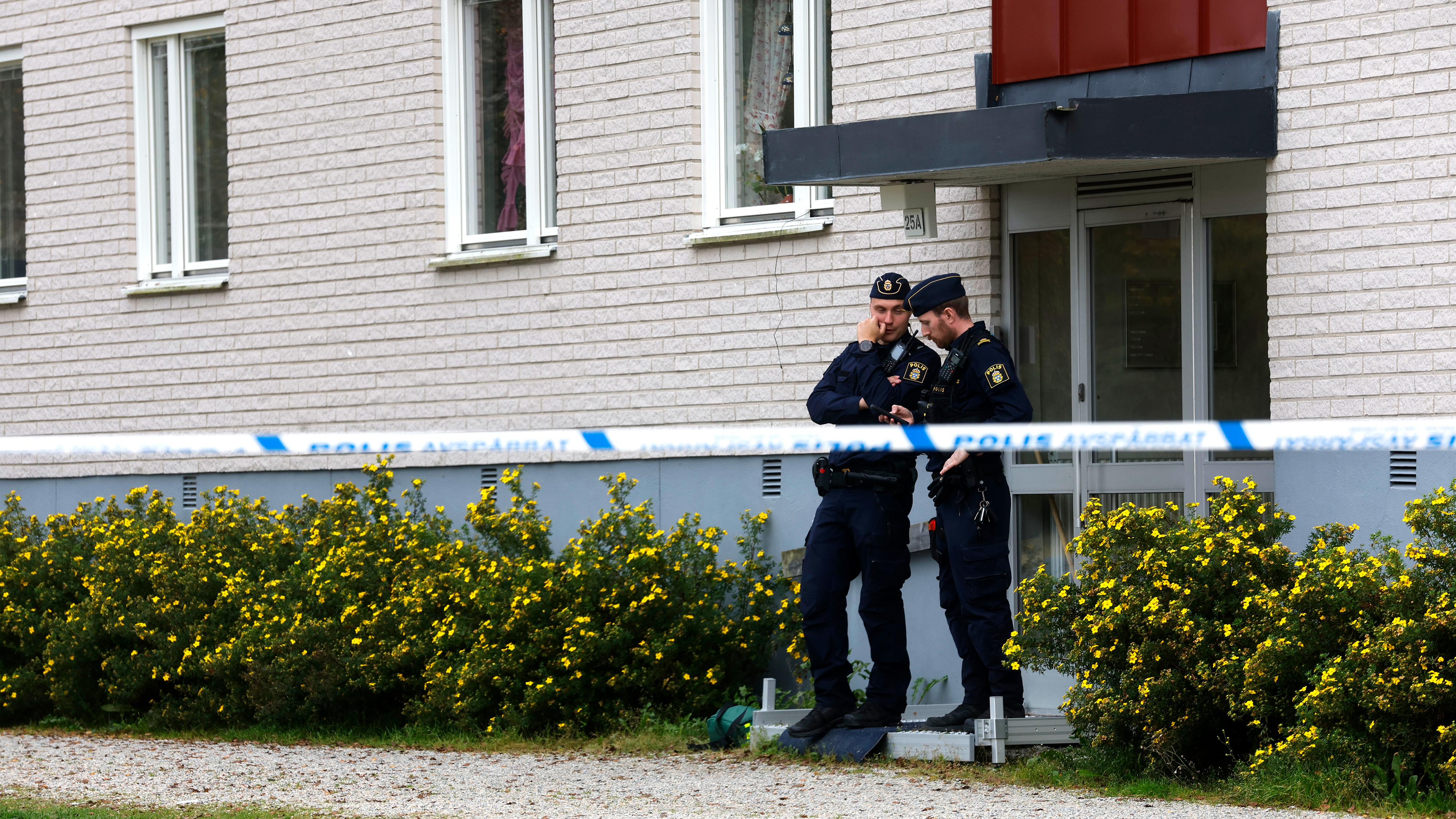 Två poliser står och pratar utanför en byggnadsentré bakom polistejp, med buskar och fönster i bakgrunden.