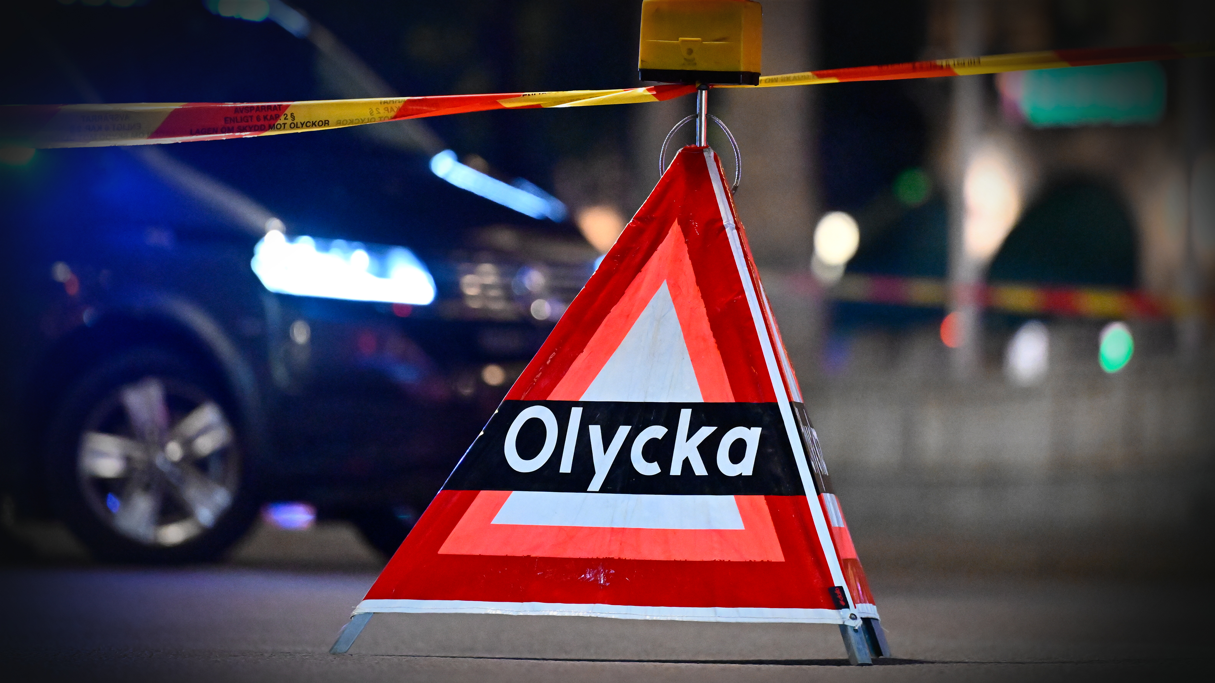 En trekantig varningsskylt med texten "Olycka" står framför en bil och polistejp på natten.