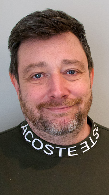 Magnus Carlström med kort brunt hår och skägg ler svagt. Han har på sig en grön tröja med vit och svart krage där det står "LACOSTE". Bakgrunden är ljus.