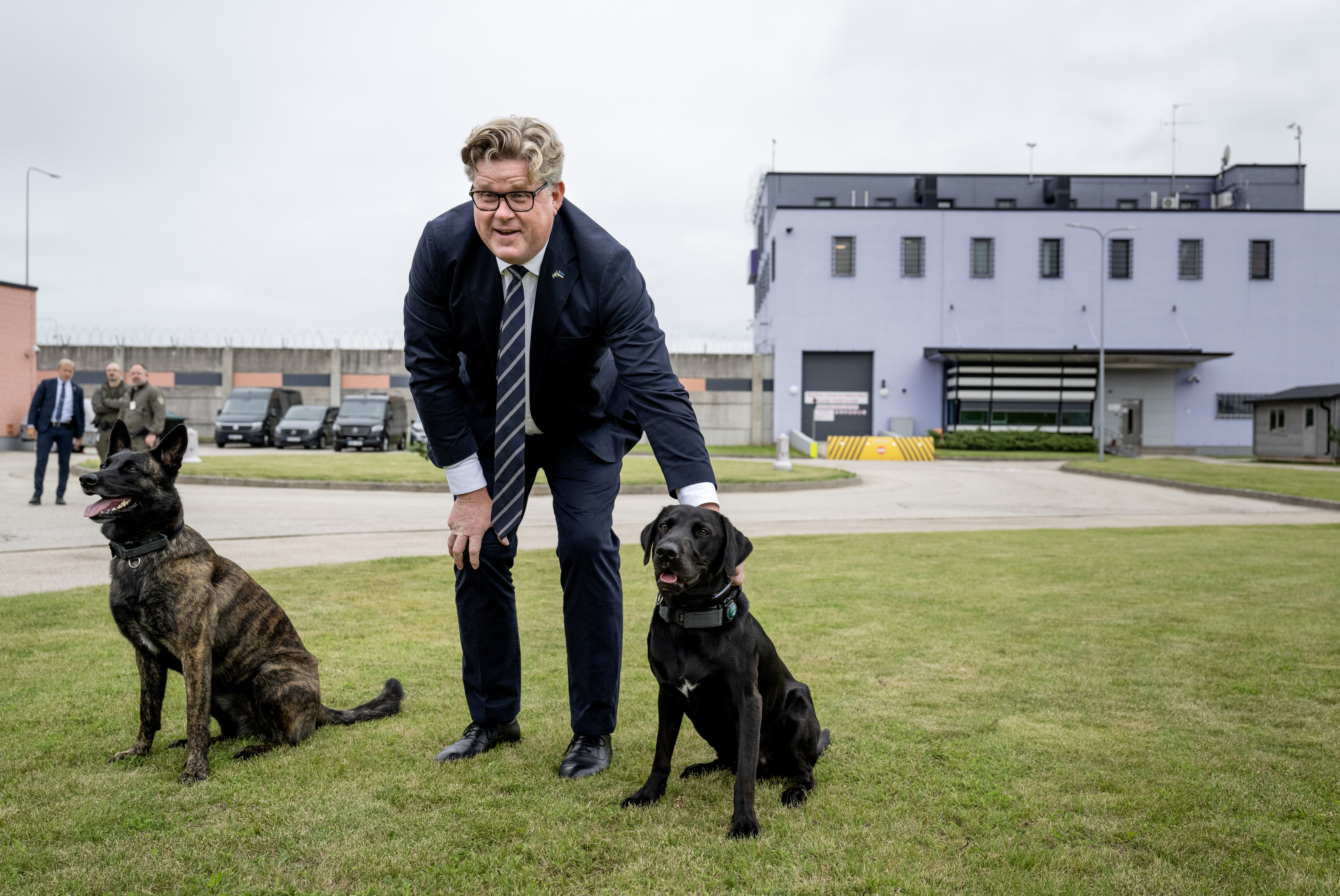 Gunnar Strömmer i kostym står på gräs och klappar en svart hund med halsband. En annan hund sitter bredvid. Flera personer och fordon syns vid en stor, ljuslila byggnad i bakgrunden.