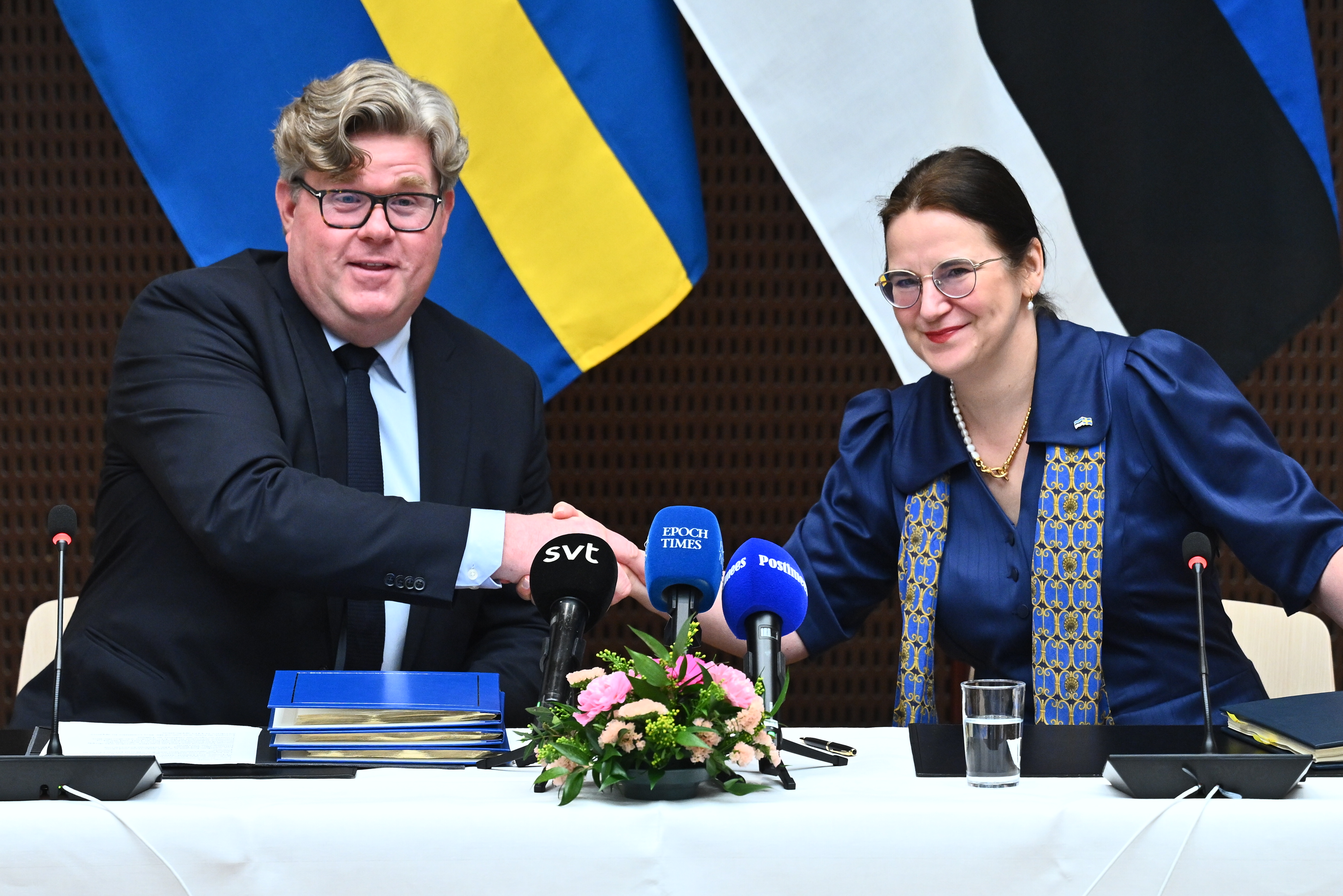 Gunnar Strömmer (M) och Liisa-Ly Pakosta sitter vid ett bord och tar i hand framför svenska och estniska flaggor, med mikrofoner, dokument och blommor på bordet.