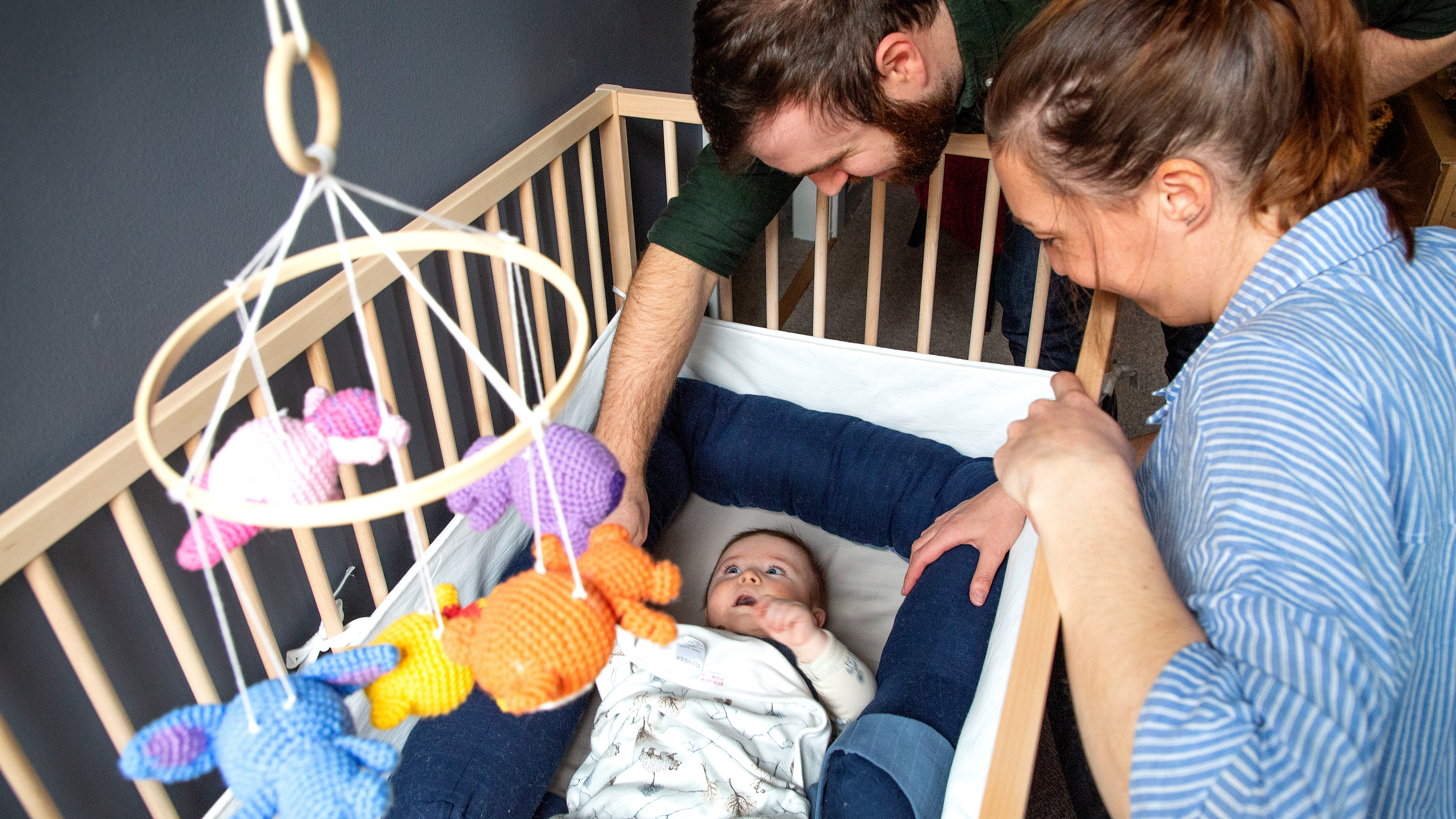 Två vuxna lutar sig över en spjälsäng och tittar på en baby som ligger där. Ett mobil med stickade djur hänger ovanför sängen.
