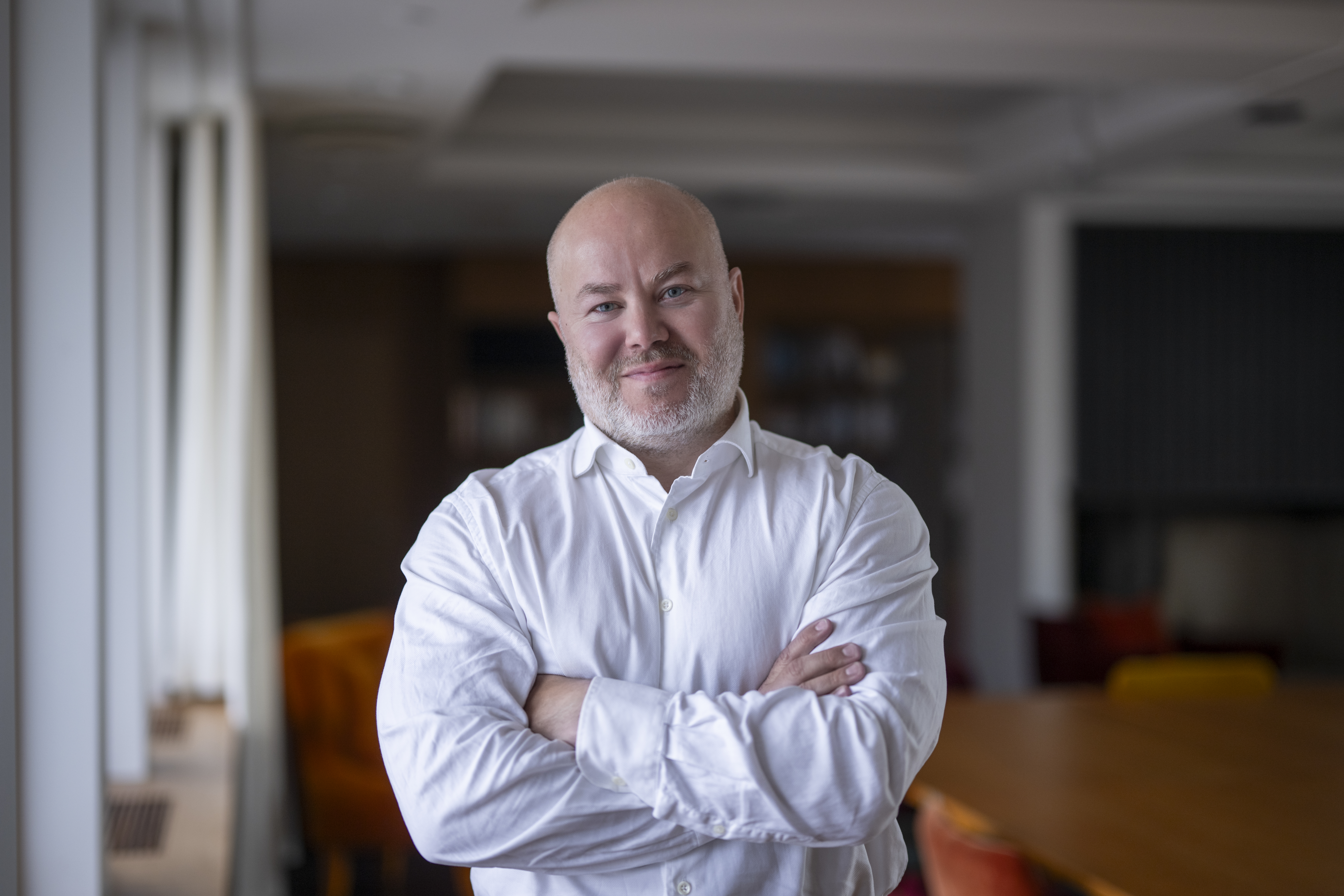 En skallig man med skägg, klädd i vit skjorta, står med armarna i kors i ett kontor eller konferensrum.