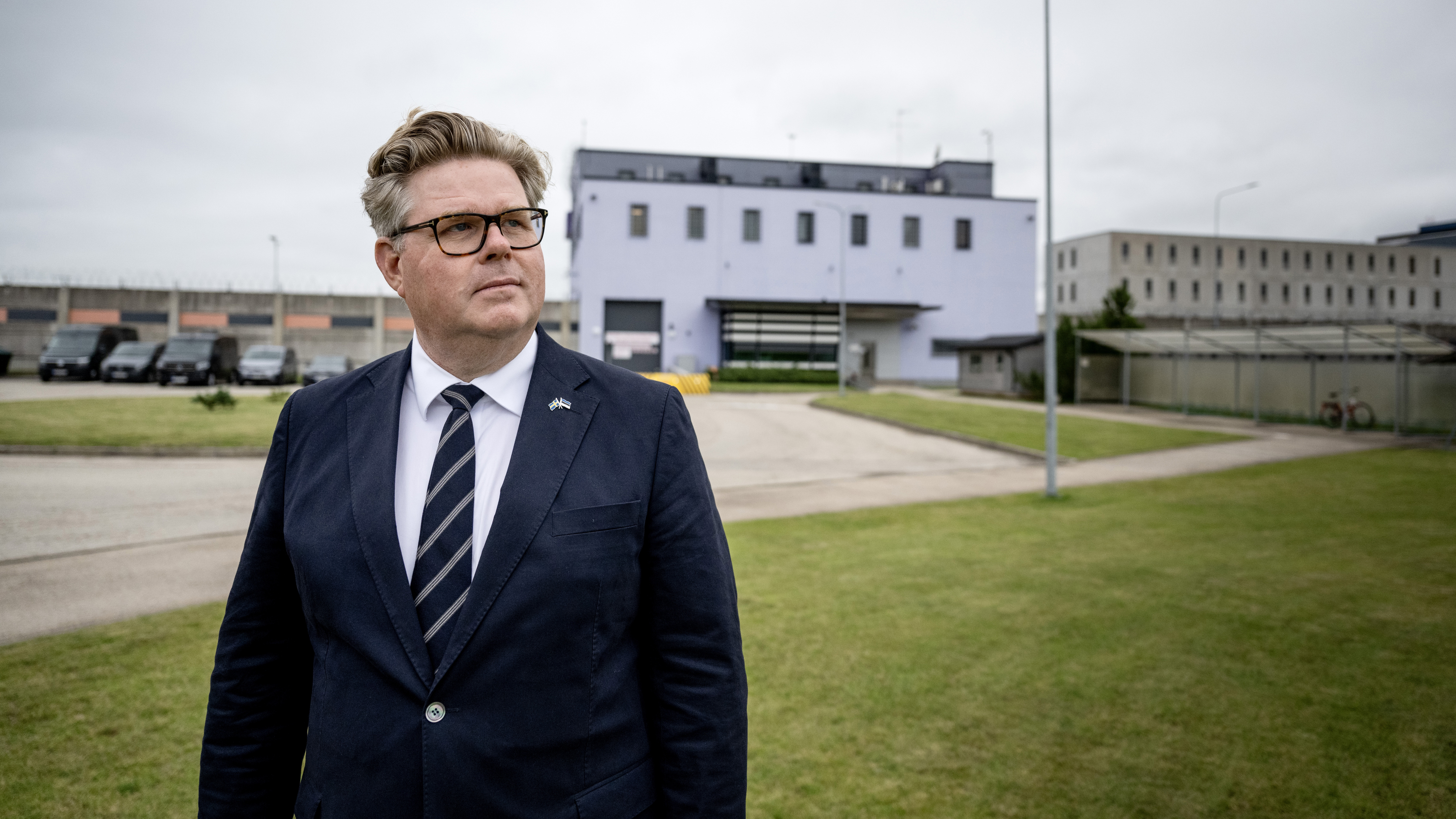 Gunnar Strömmer i kostym och slips står på gräs framför en stor byggnad under en mulen himmel.