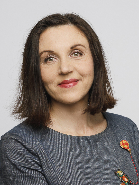 Anna Markina med brunt axellångt hår i grå topp och röd brosch, vänd mot kameran och ler mot en ljus bakgrund.