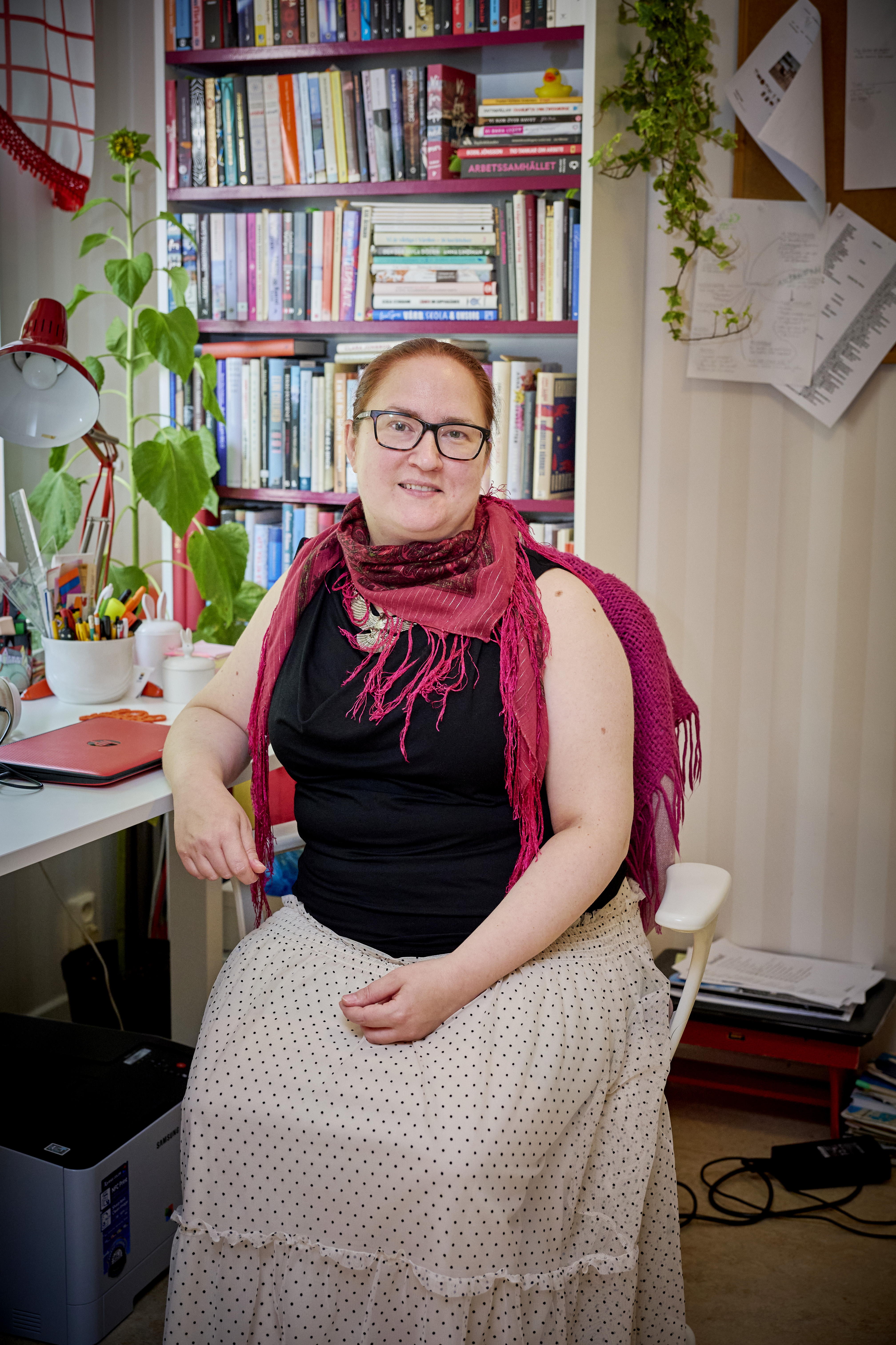 En kvinna med glasögon och röd scarf sitter på en vit stol i ett hemmakontor med bokhylla, skrivbord och växter bakom.