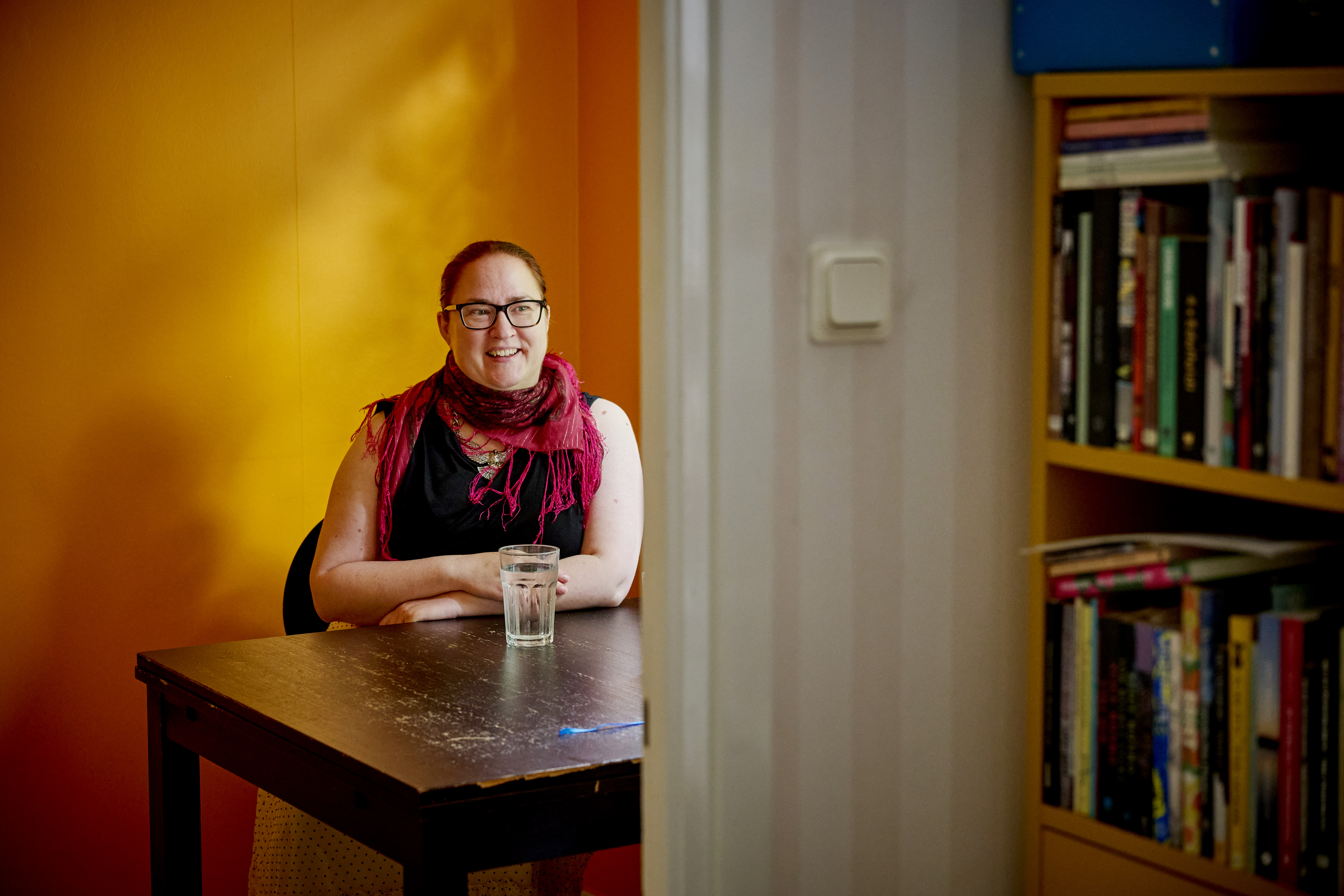 En person med glasögon och röd scarf sitter vid ett bord med ett vattenglas i ett rum med orange väggar och en bokhylla.