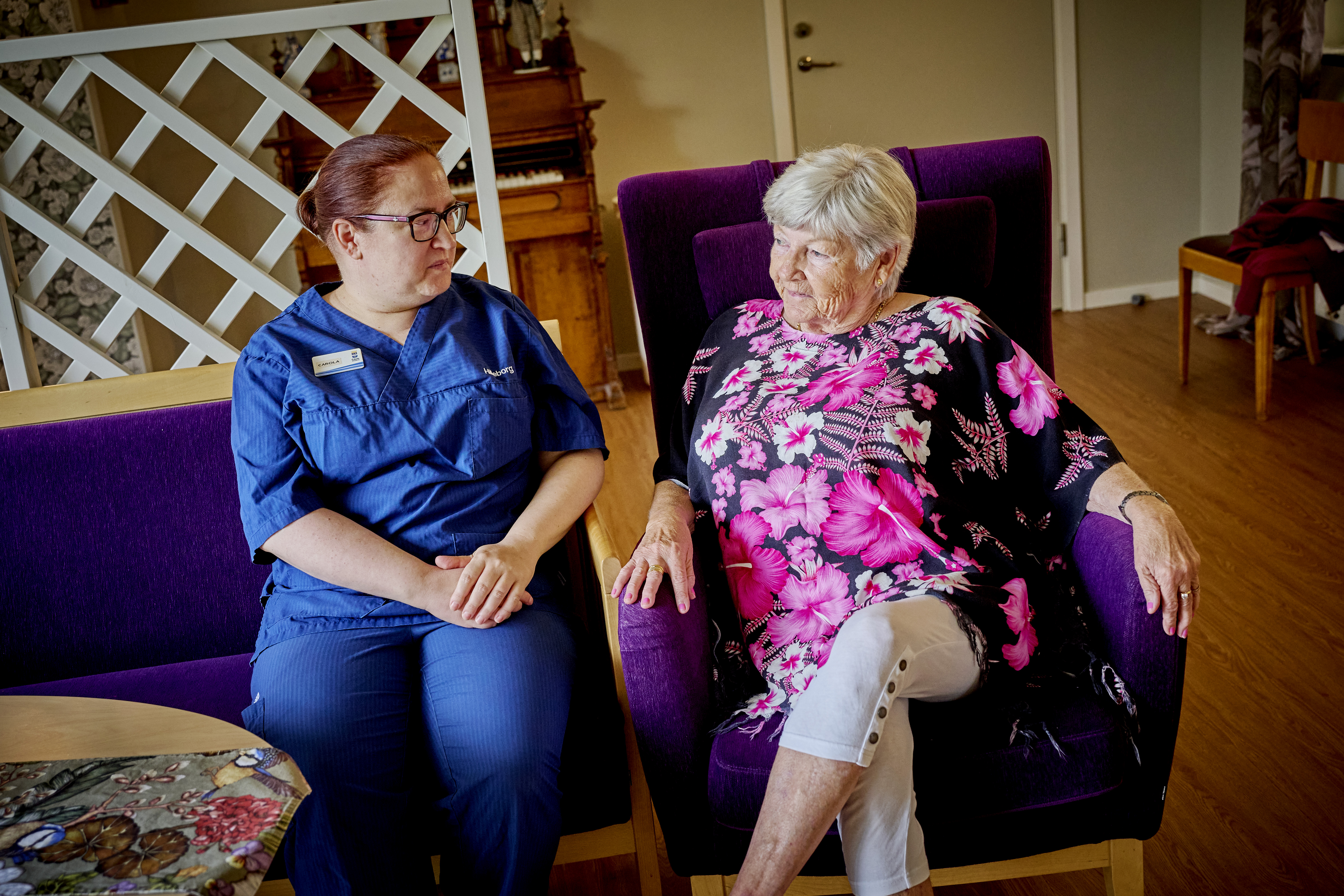 En vårdpersonal i blå arbetskläder sitter bredvid en äldre kvinna i blommig topp på en stol och samtalar på ett vårdboende.