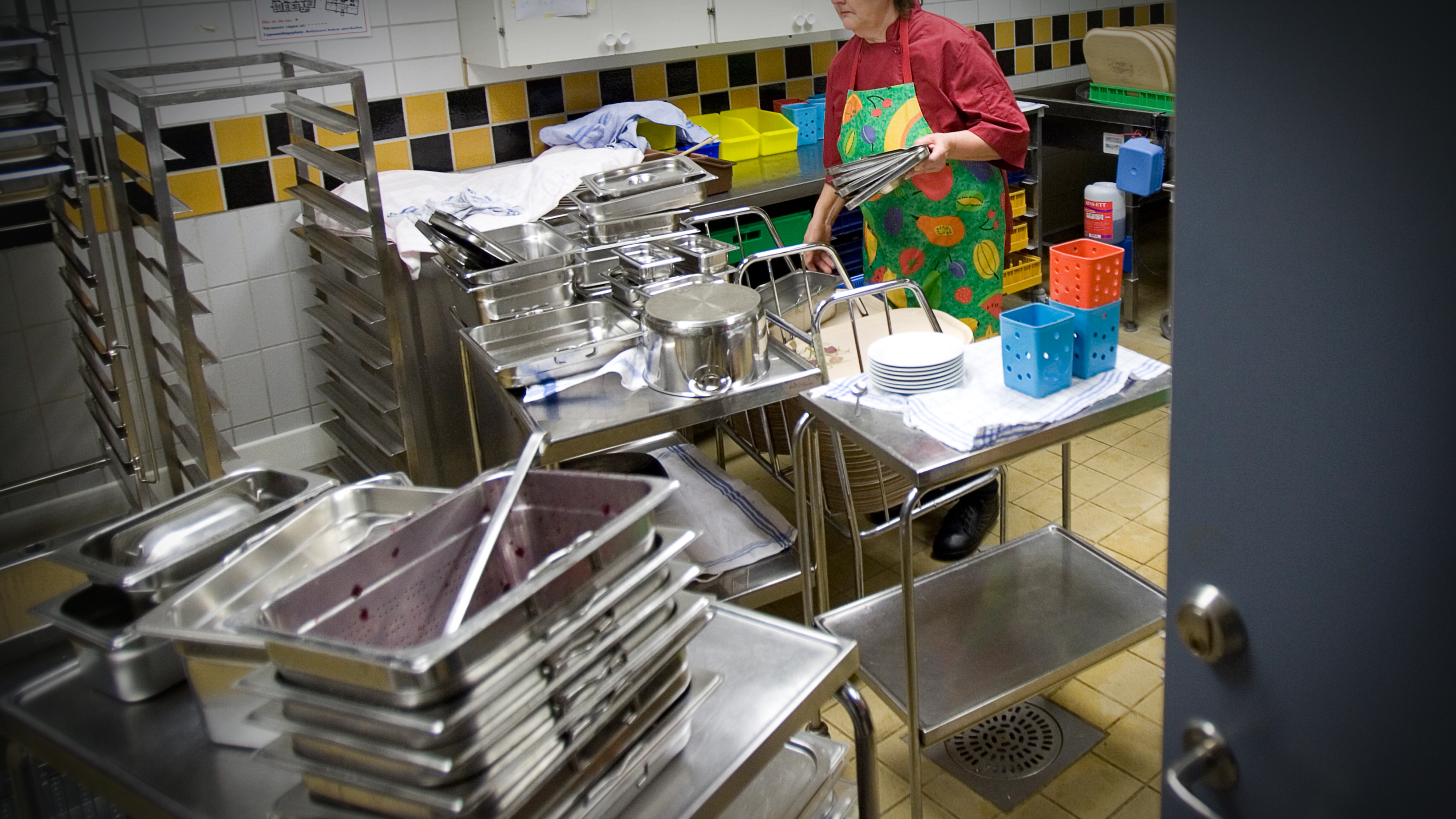 En person står i ett storkök omgiven av staplar med metallbrickor, kastruller och tallrikar och förbereder sig på att städa upp.