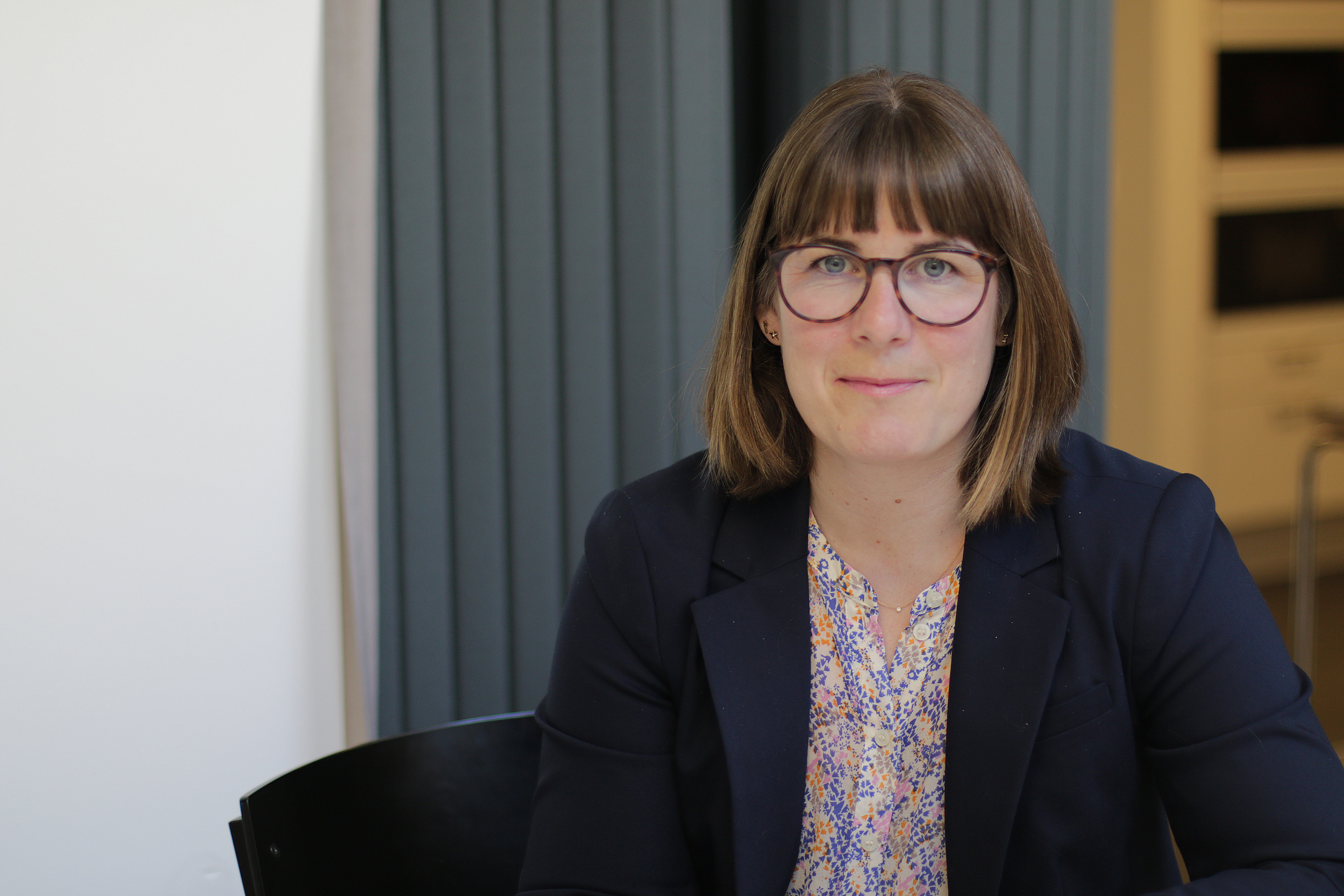 Lisa Gezelius med rakt brunt hår och glasögon, klädd i mörkblå kavaj och mönstrad blus, sitter vid ett bord inomhus.