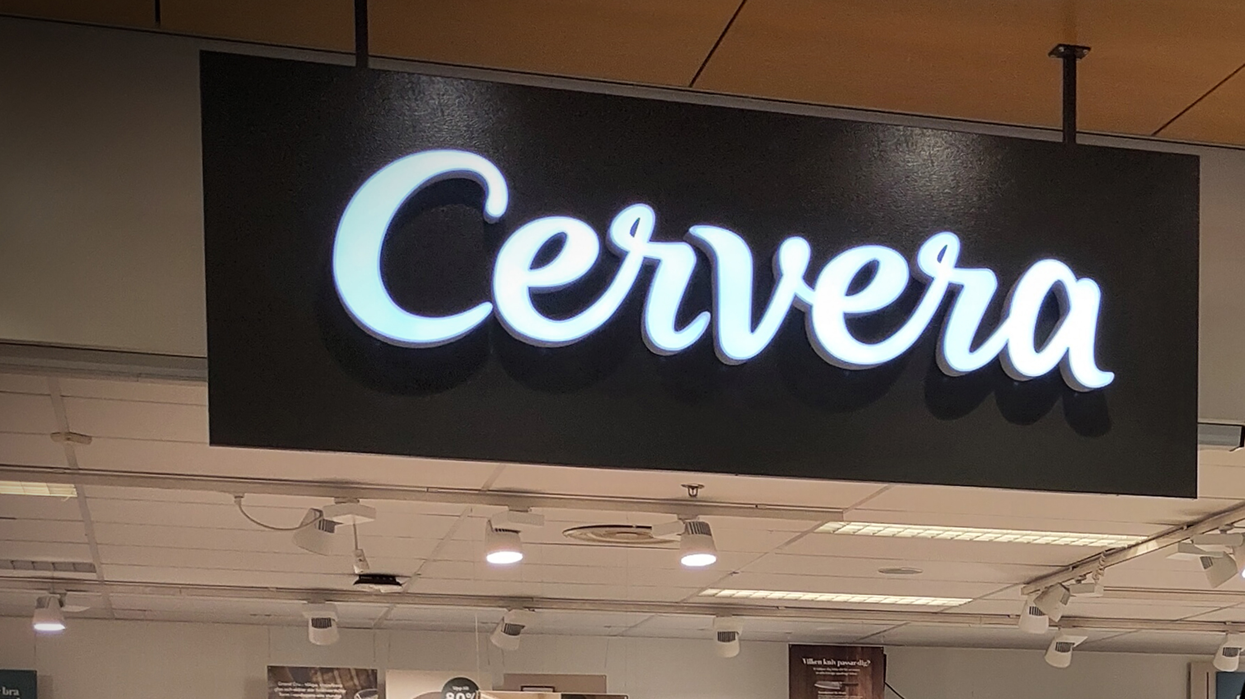 En svart rektangulär skylt med ordet "Cervera" i vita skrivna bokstäver, hängande från ett innertak i en inomhusmiljö.
