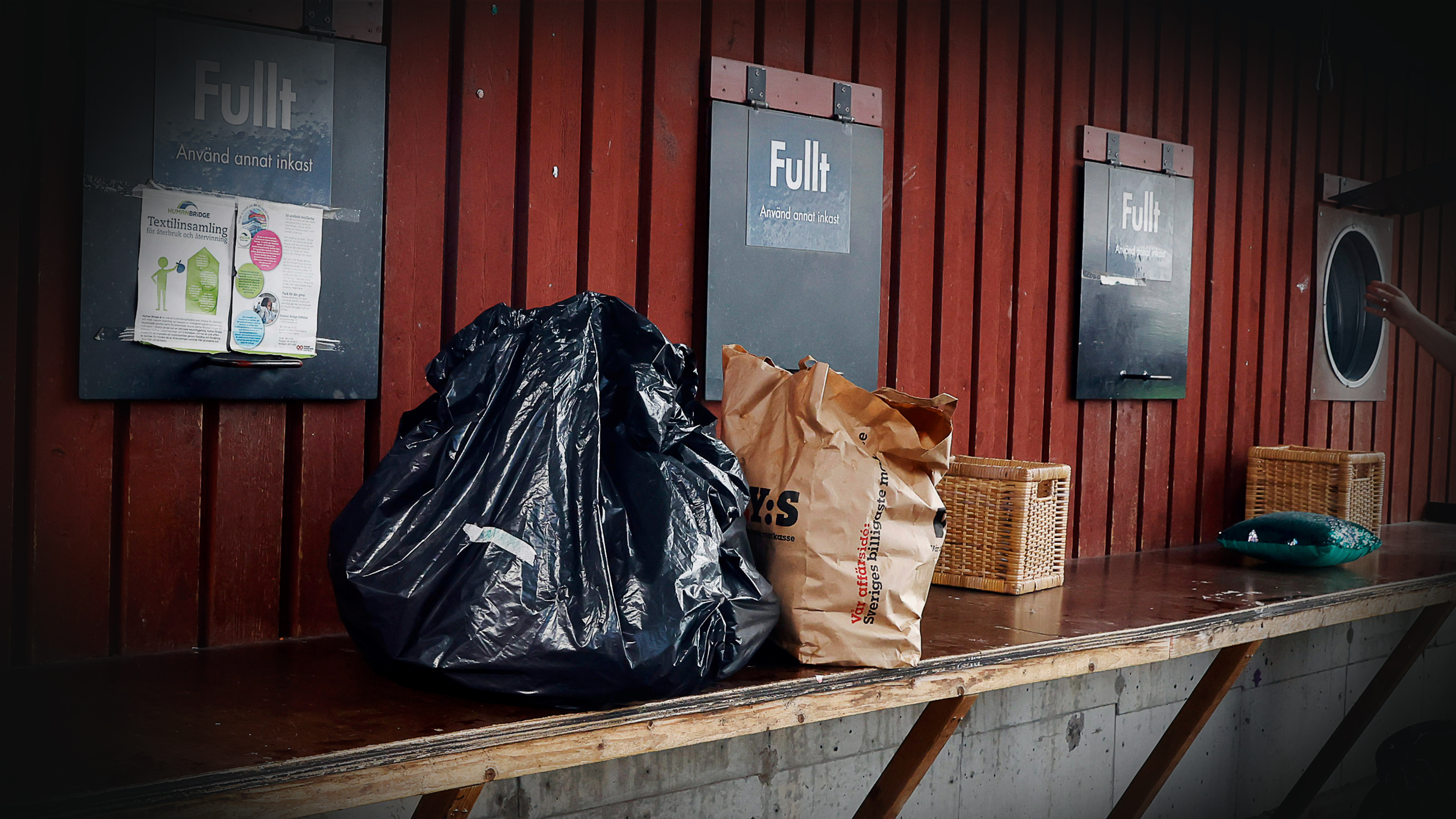 En svart plastpåse och en brun papperspåse placeras på en bänk framför märkta behållare "helt" På återvinningsstationen.