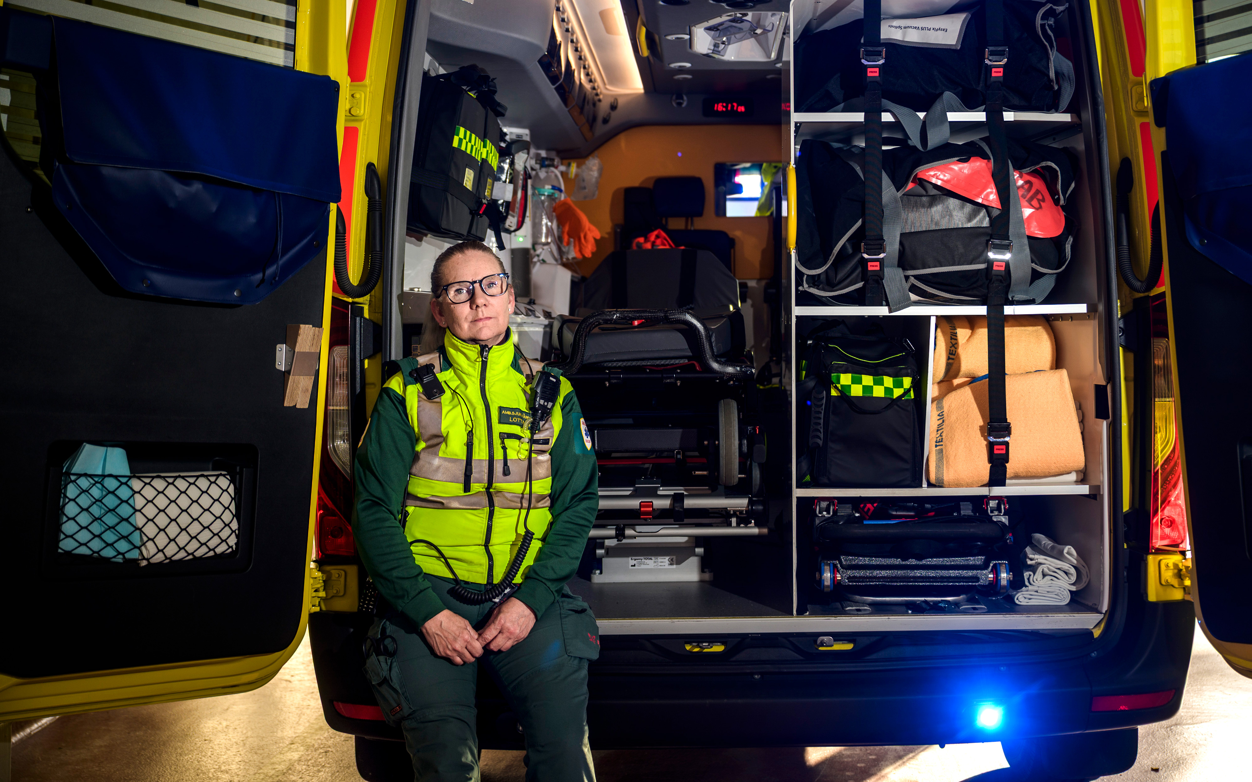 En ambulanspersonal i uniform sitter vid den öppna bakdörren till en ambulans med medicinsk utrustning och material runt sig.