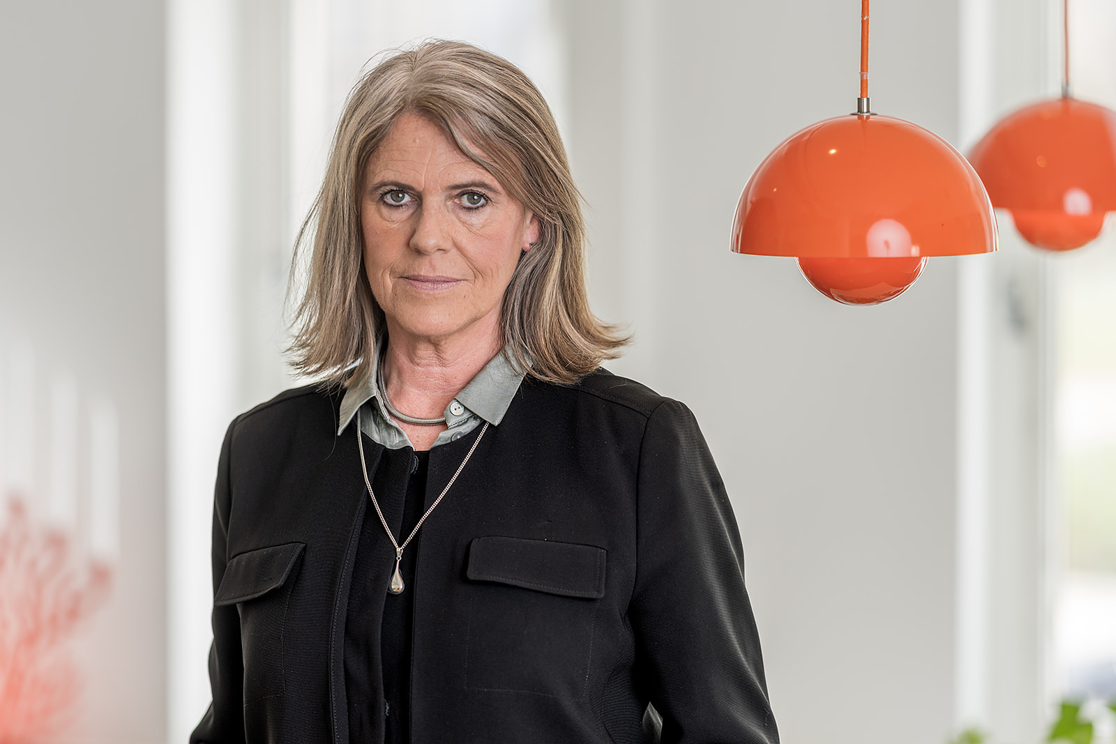 En kvinna med axellångt grått hår står inomhus med en svart jacka över en skjorta. Två orange lampor syns i bakgrunden.