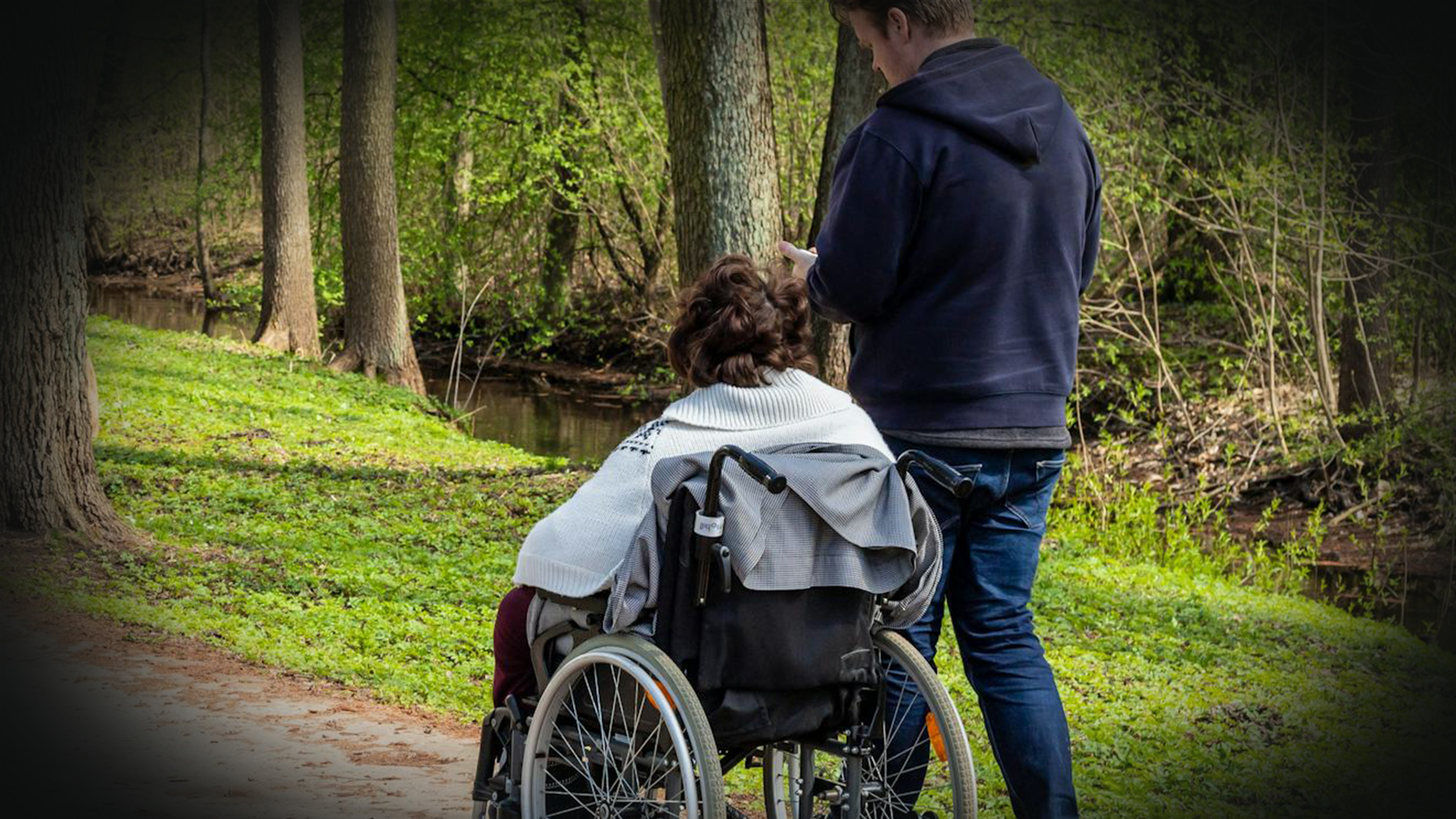 En person i rullstol får sällskap av en annan person som går bredvid på en stig i ett skogsområde.