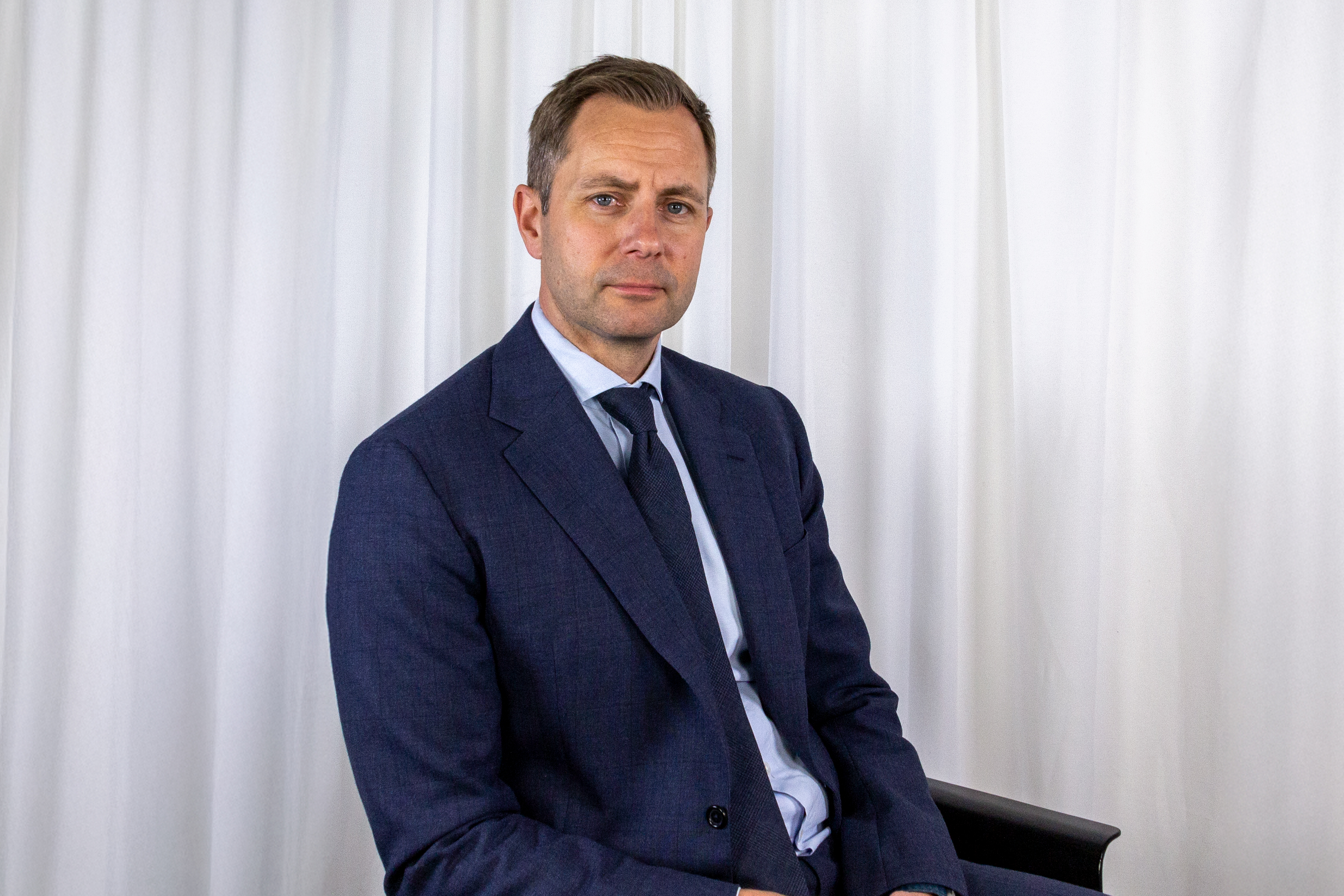 En man i kostym och slips sitter på en stol framför ett vitt draperi och tittar mot kameran med neutral min.