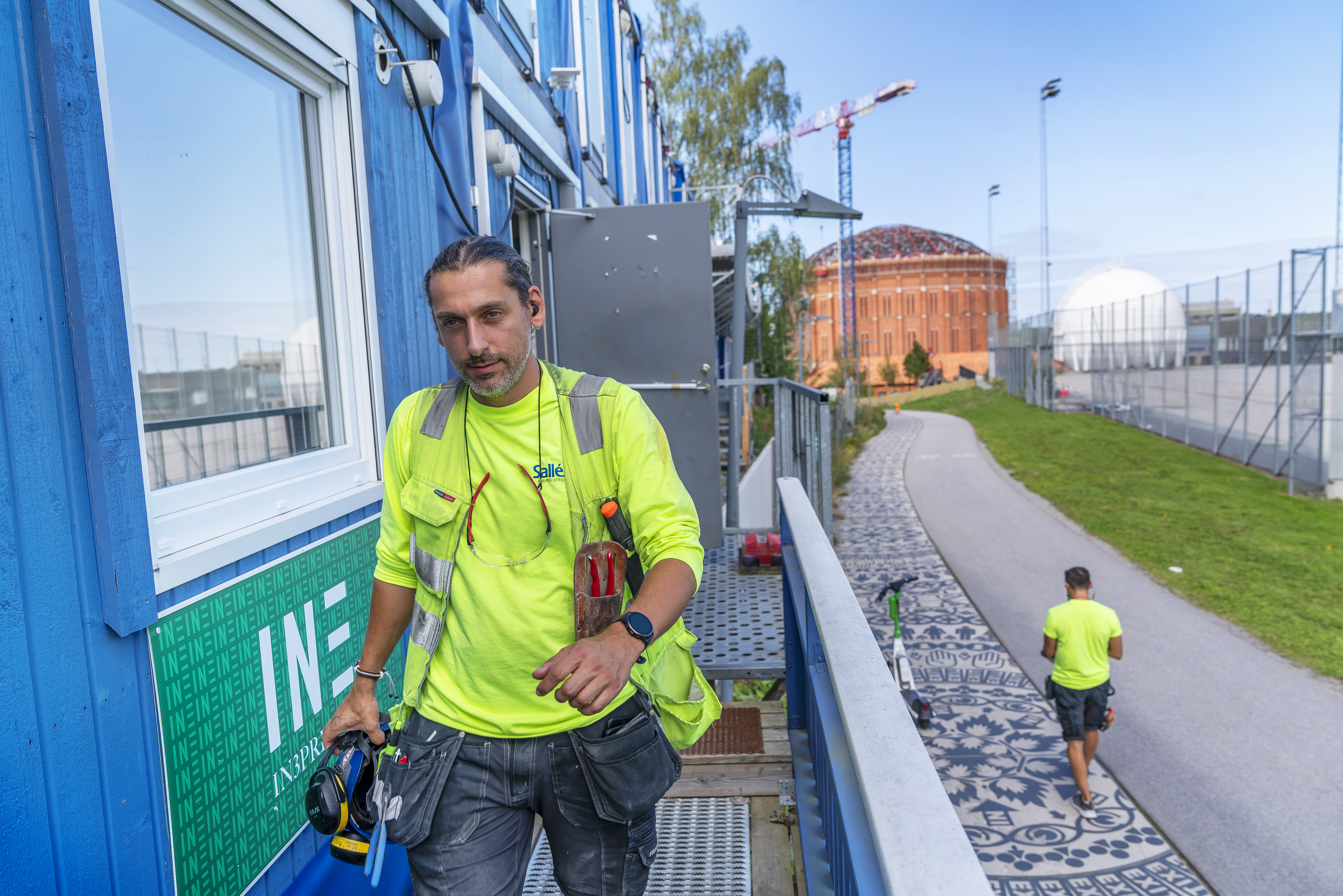 Två byggnadsarbetare har på sig gula skjortor; en går på en mönstrad gång, den andra står på en blå trappa med verktyg.