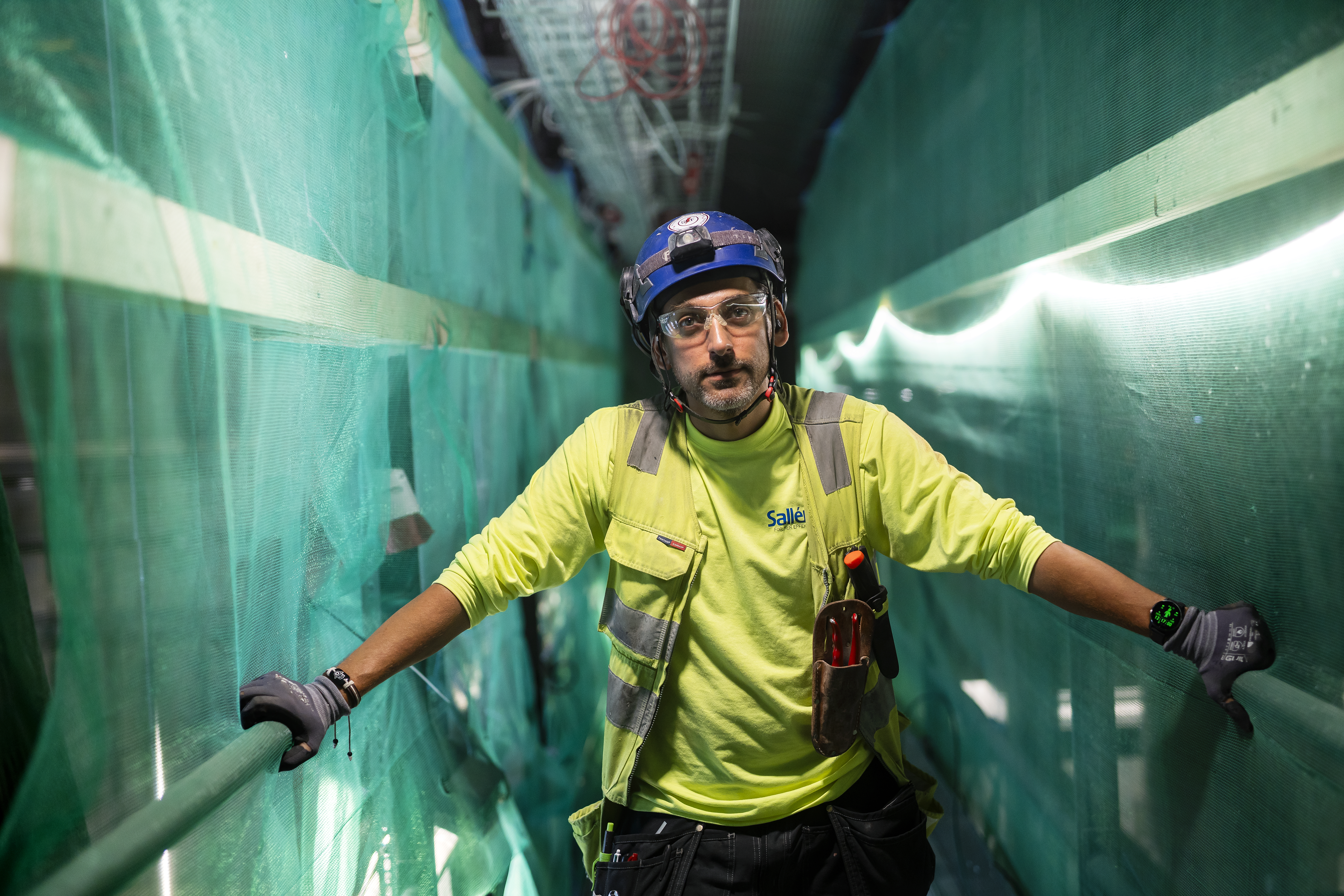 En byggarbetare med neonskjorta, skyddsväst, handskar och hjälm står i en smal gång med grönt nät och tittar mot kameran.