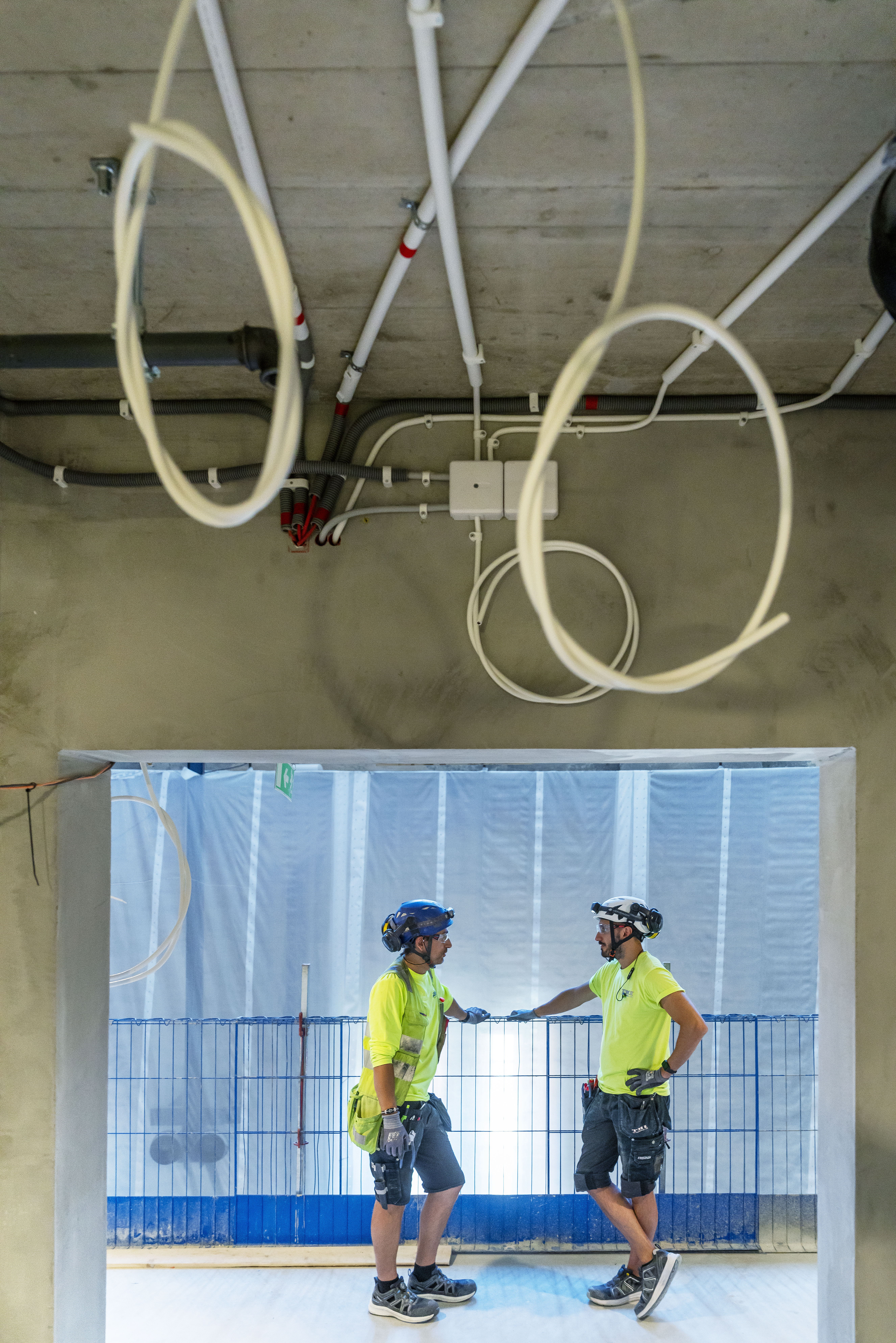 Två byggnadsarbetare med skyddsutrustning står och pratar under rör och ledningar i ett ofärdigt byggnadsutrymme.