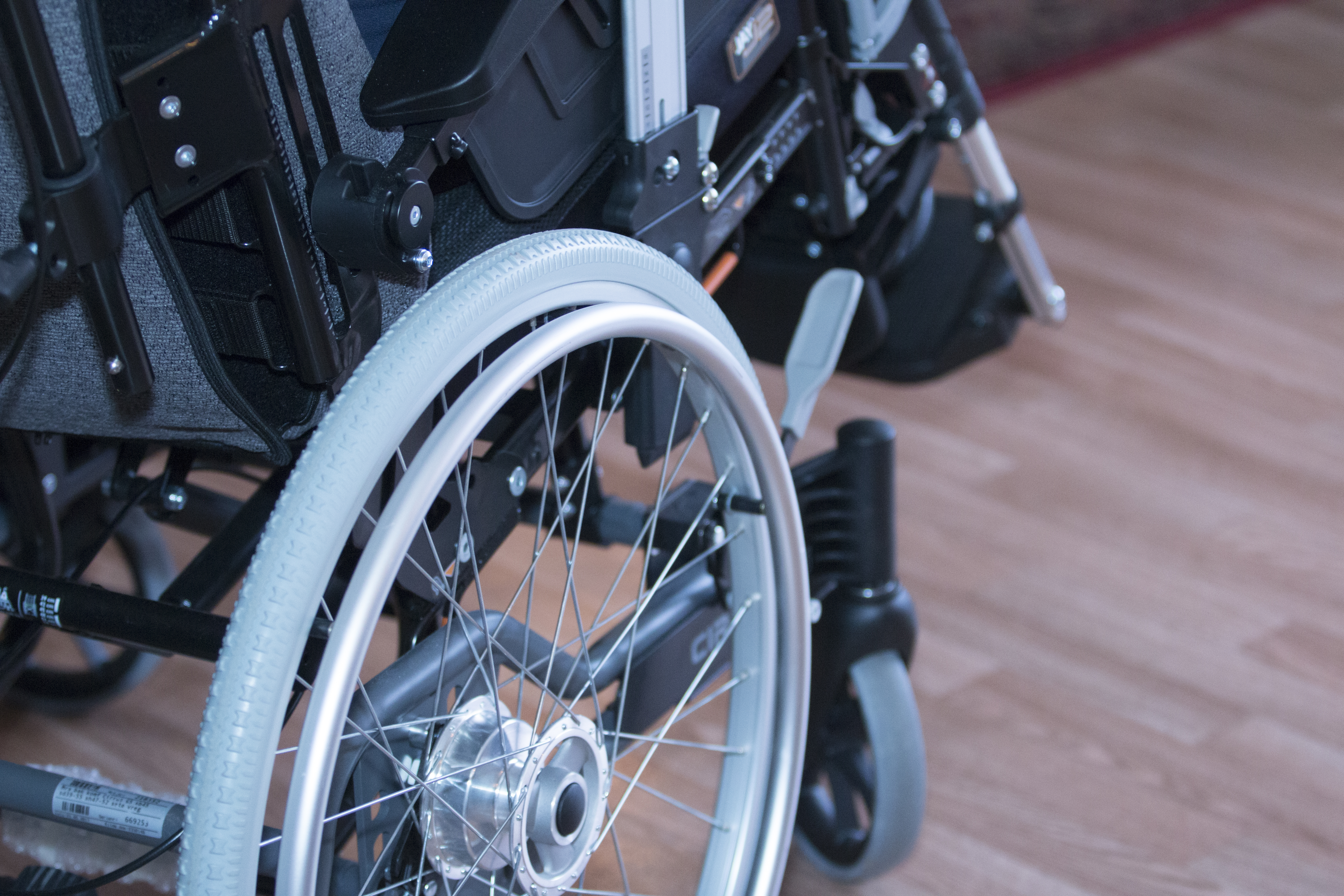 Närbild av en rullstol på ett trägolv, där hjul, ram och en del av sitsen syns.
