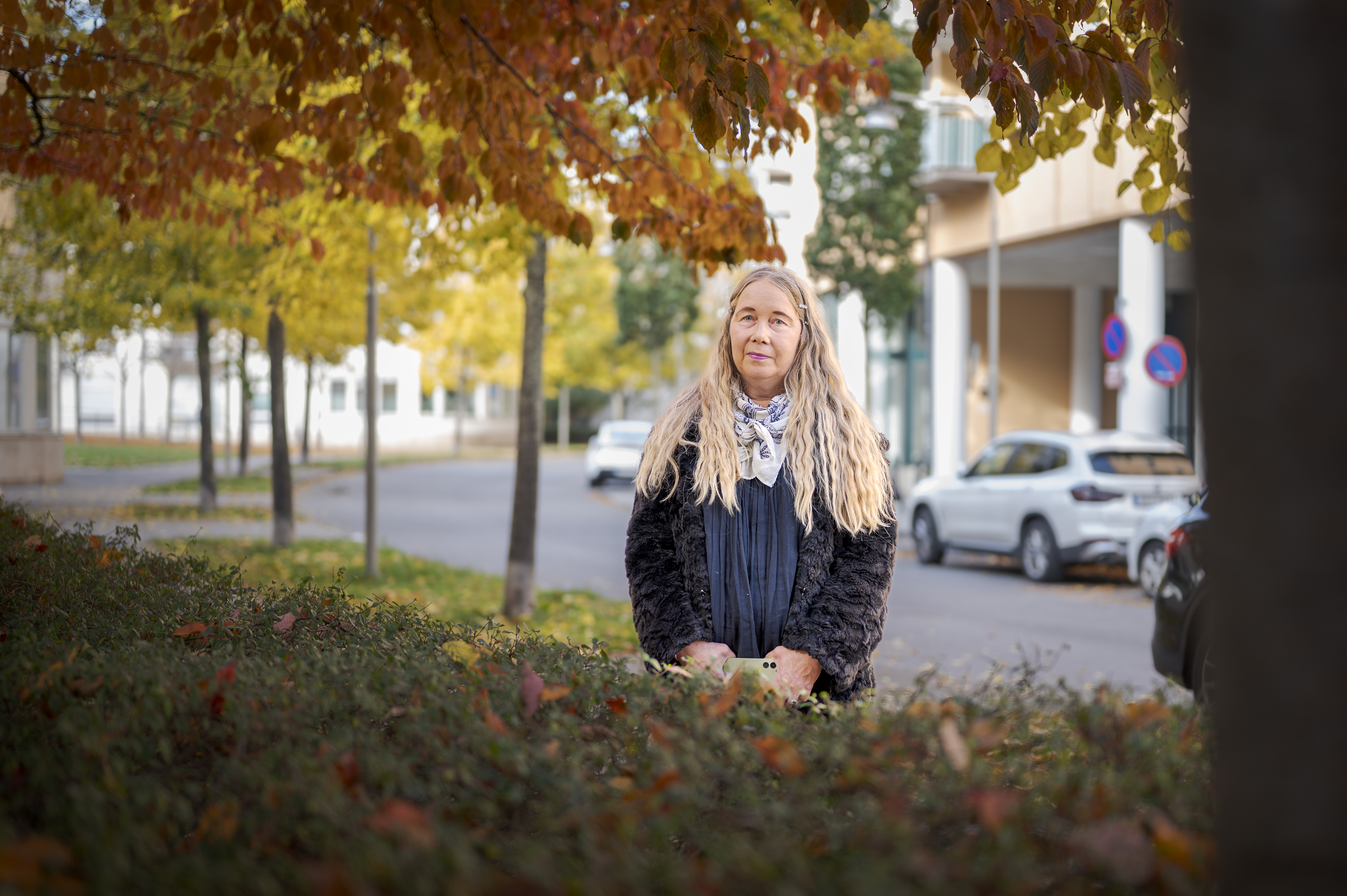 Maria Tornefjell med långt blont hår står utomhus nära en häck, under träd, med parkerade bilar och byggnader i bakgrunden.