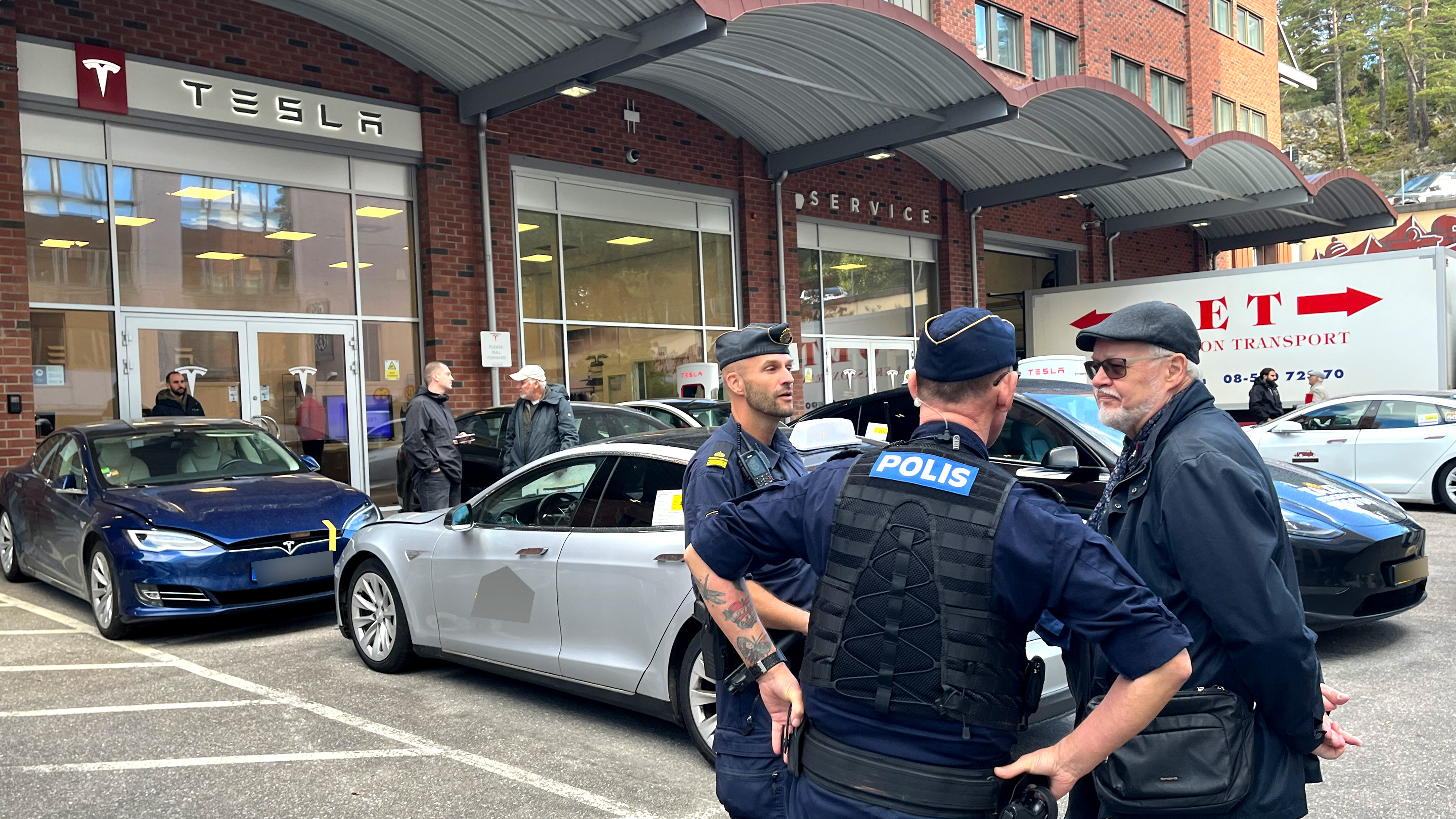 Poliser står och pratar utanför en Tesla-verkstad med flera bilar och personer i bakgrunden.