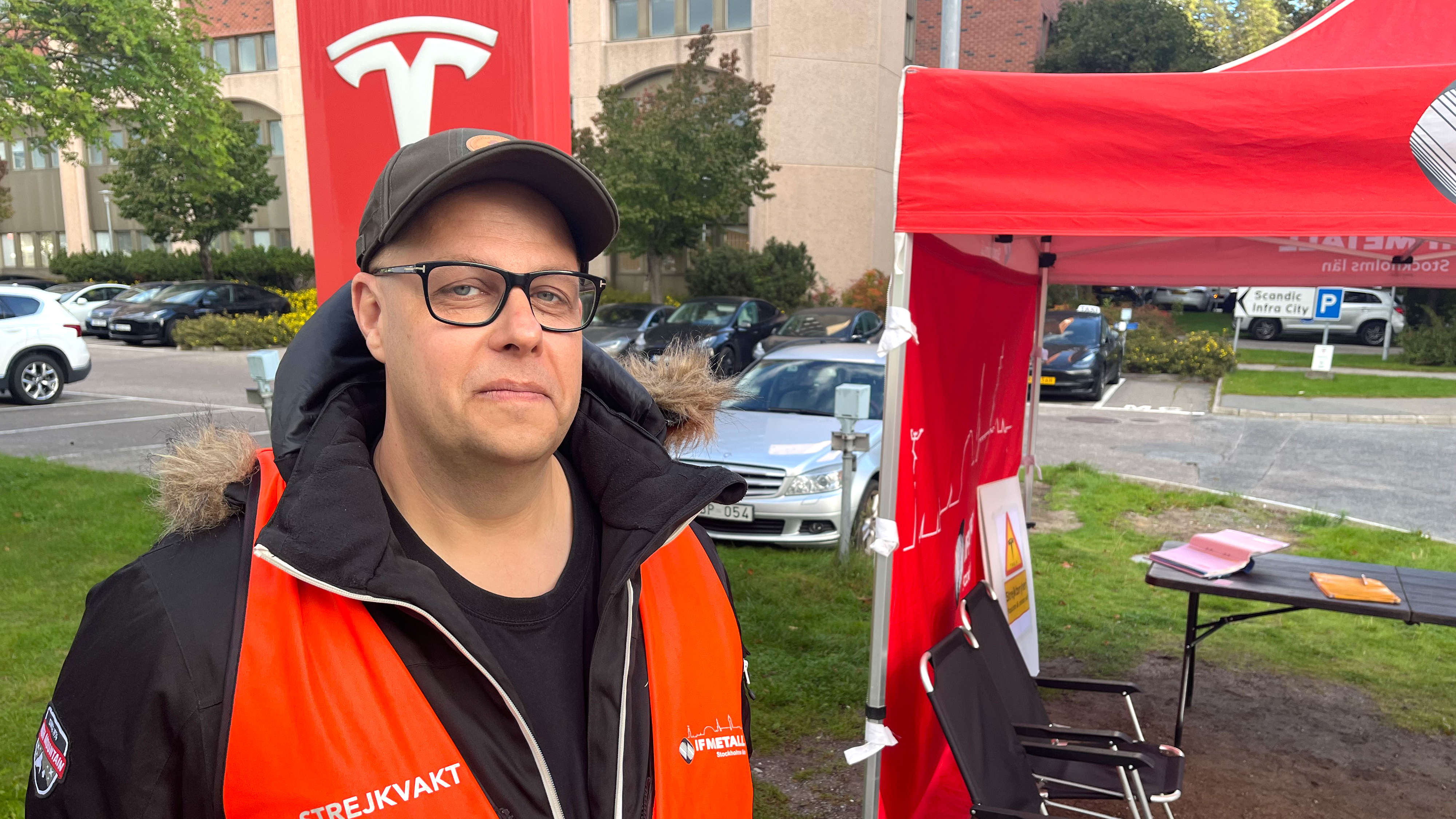 En person med glasögon och orange väst står utomhus nära en Tesla-banner och ett rött tält på en parkering.