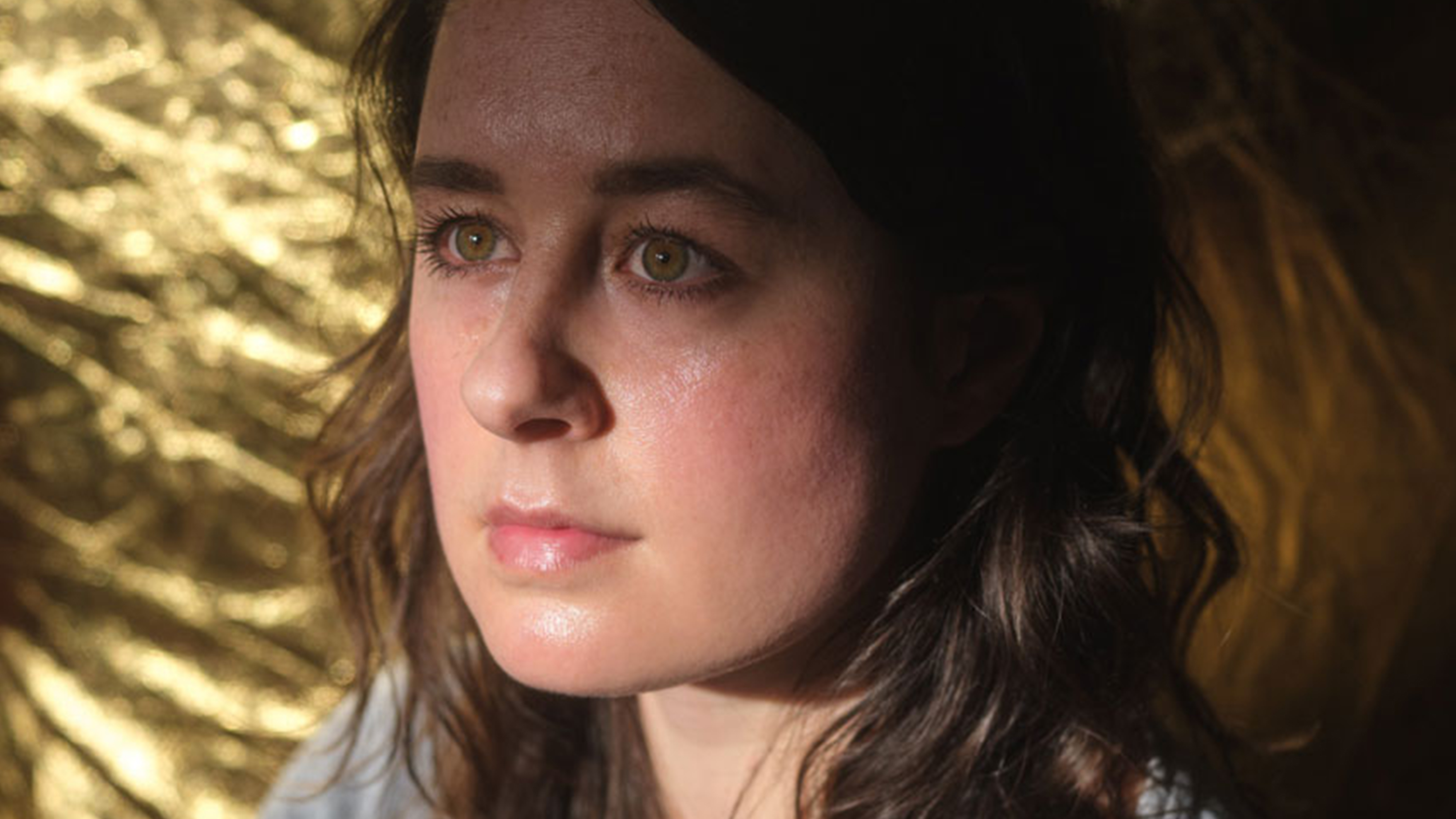 En kvinna med vågigt brunt hår tittar lite uppåt, med mjukt ljus och en guldfärgad bakgrund bakom sig.