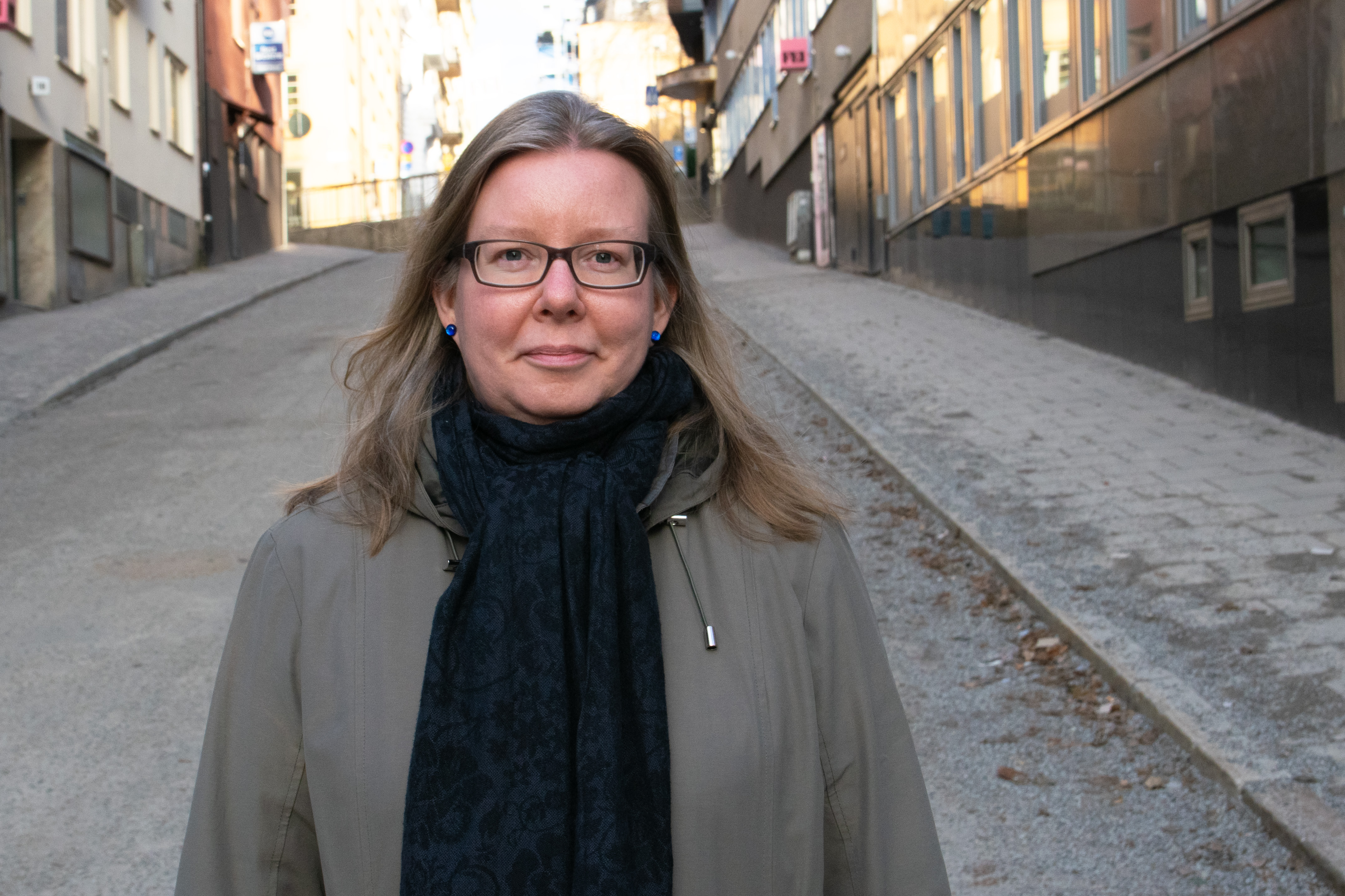 Johanna Skinnari med glasögon och långt hår står på en stadsgata som lutar uppåt, omgiven av byggnader, och har på sig kappa och halsduk.