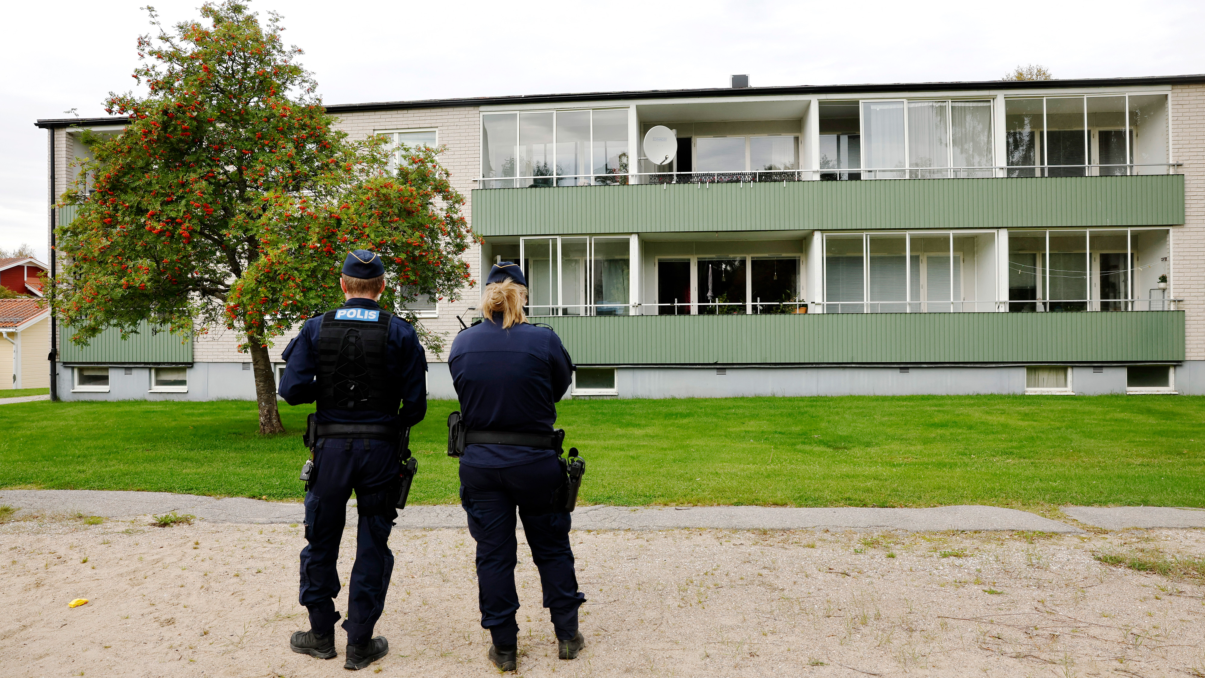 Två poliser står utanför ett bostadshus och vänder sig mot balkongerna och fönstren på en gräsyta.