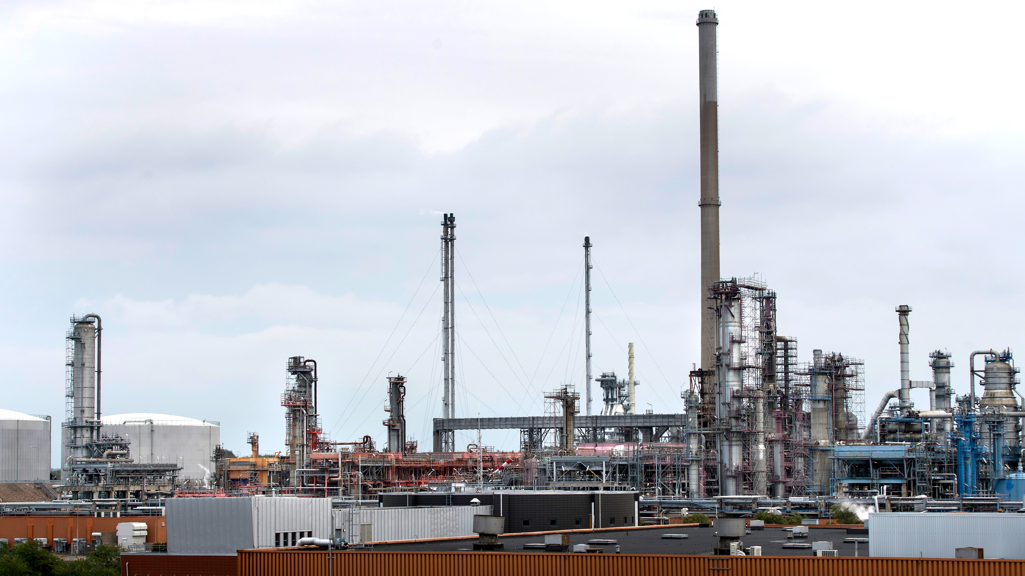 Industriellt raffinaderikomplex med höga skorstenar, rör och lagringstankar under molnig himmel. Bruna och vita byggnader syns i förgrunden.