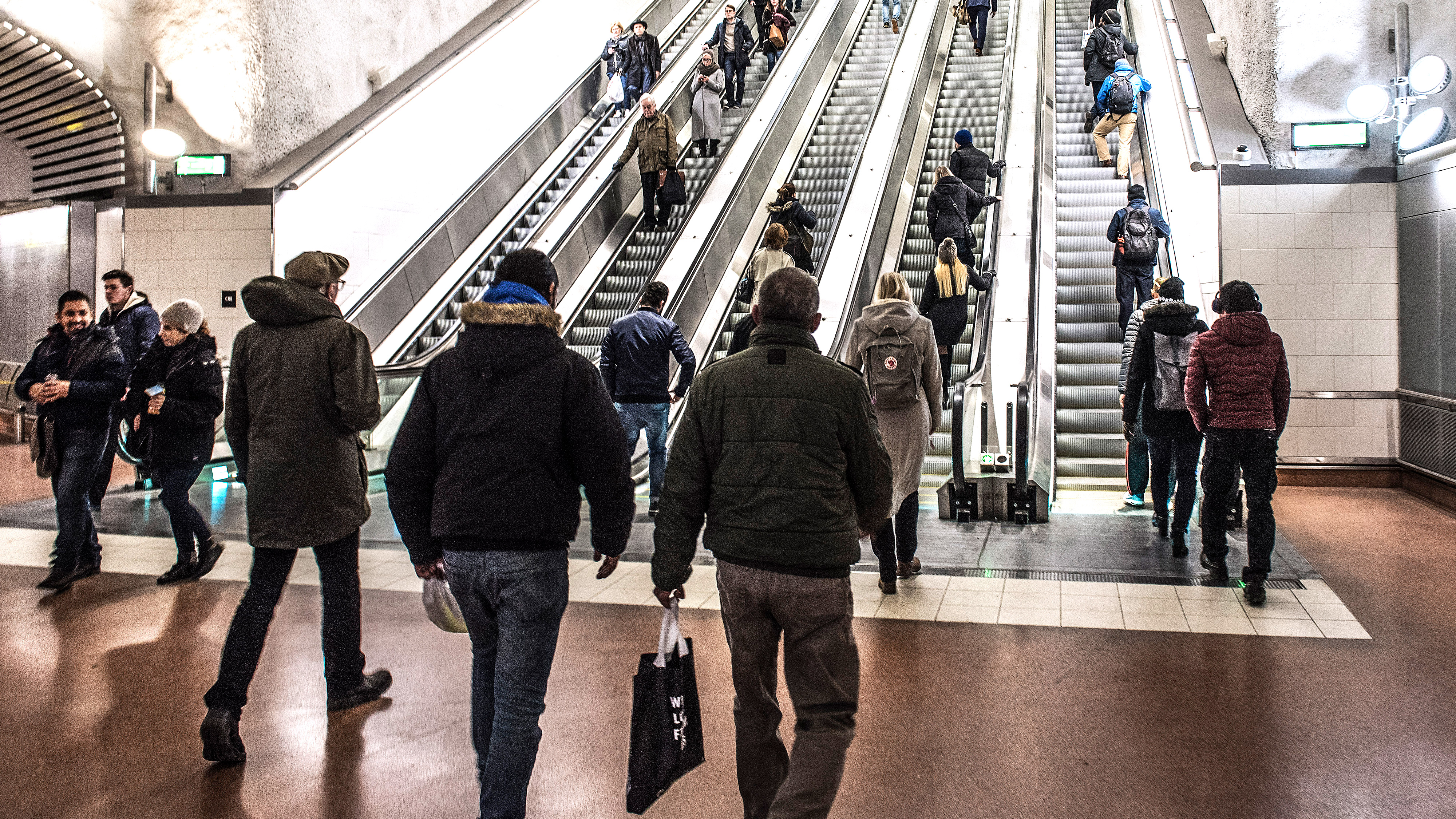 Personer går mot och åker uppför rulltrappor i en inomhusmiljö, flera bär vinterkläder.
