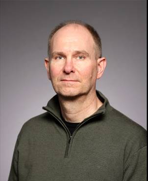 En medelålders man med kort hår och mörkgrön tröja med dragkedja står mot en grå bakgrund.