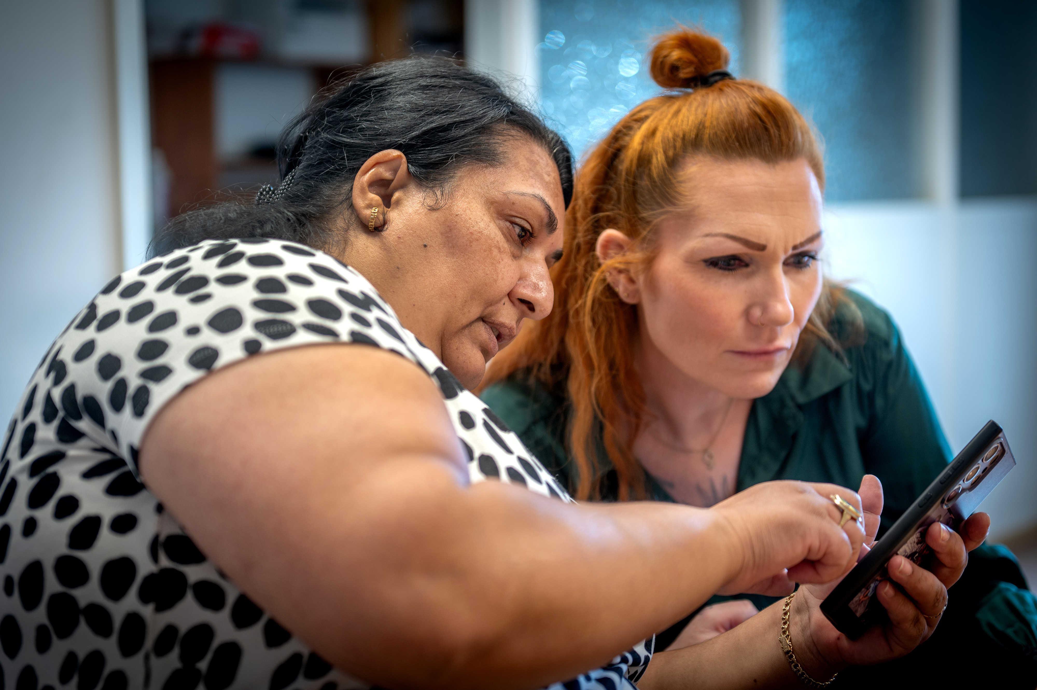 Två kvinnor sitter nära varandra och tittar på en smartphone; en av kvinnorna pekar på telefonens skärm.