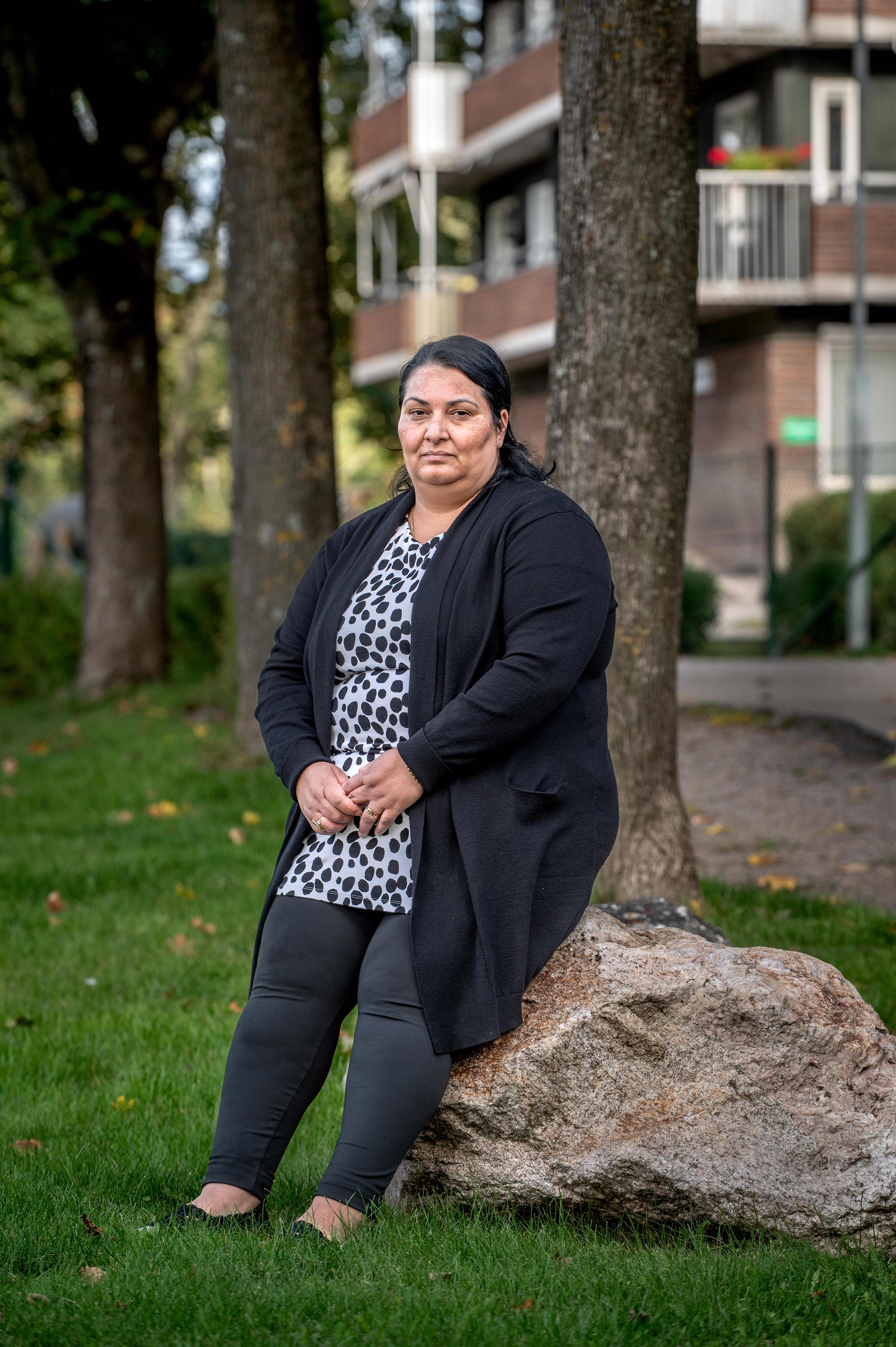 En kvinna med svart kofta och mönstrad klänning sitter på en sten i ett gräsområde, med träd och ett hyreshus i bakgrunden.