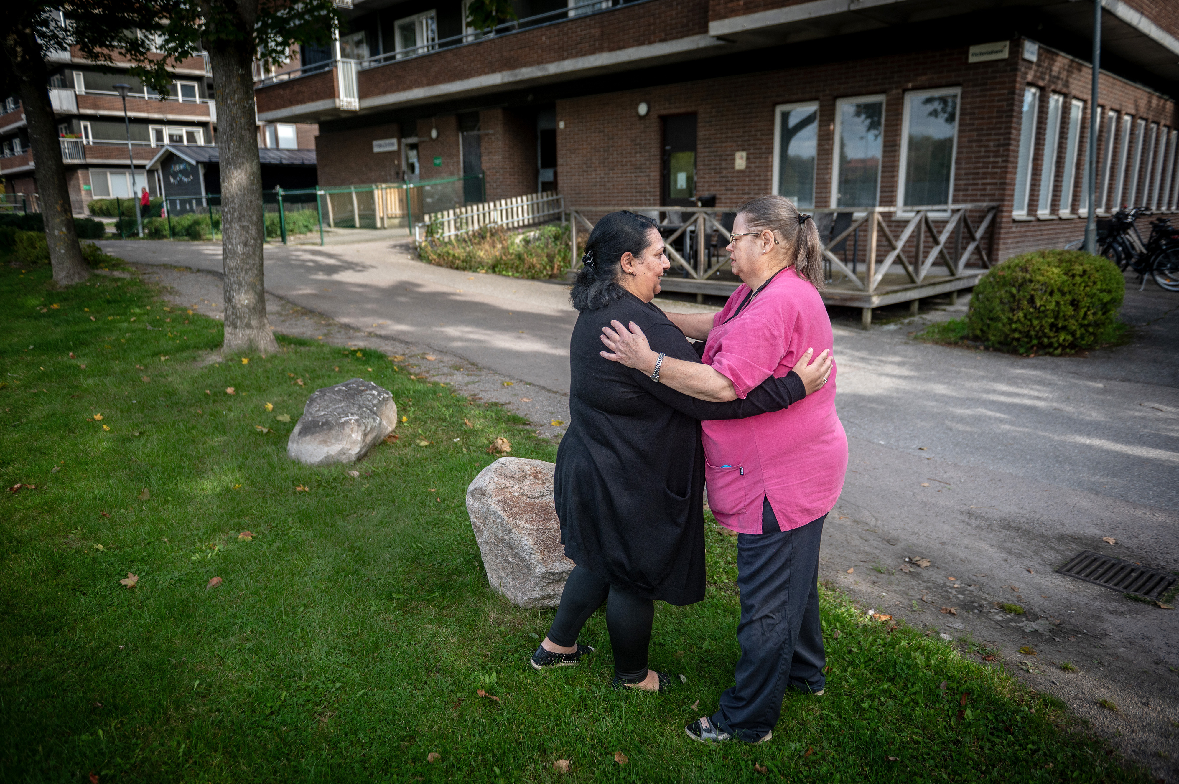 Två kvinnor står på gräset utanför ett tegelhus, vända mot varandra, rör vid varandras armar och samtalar.