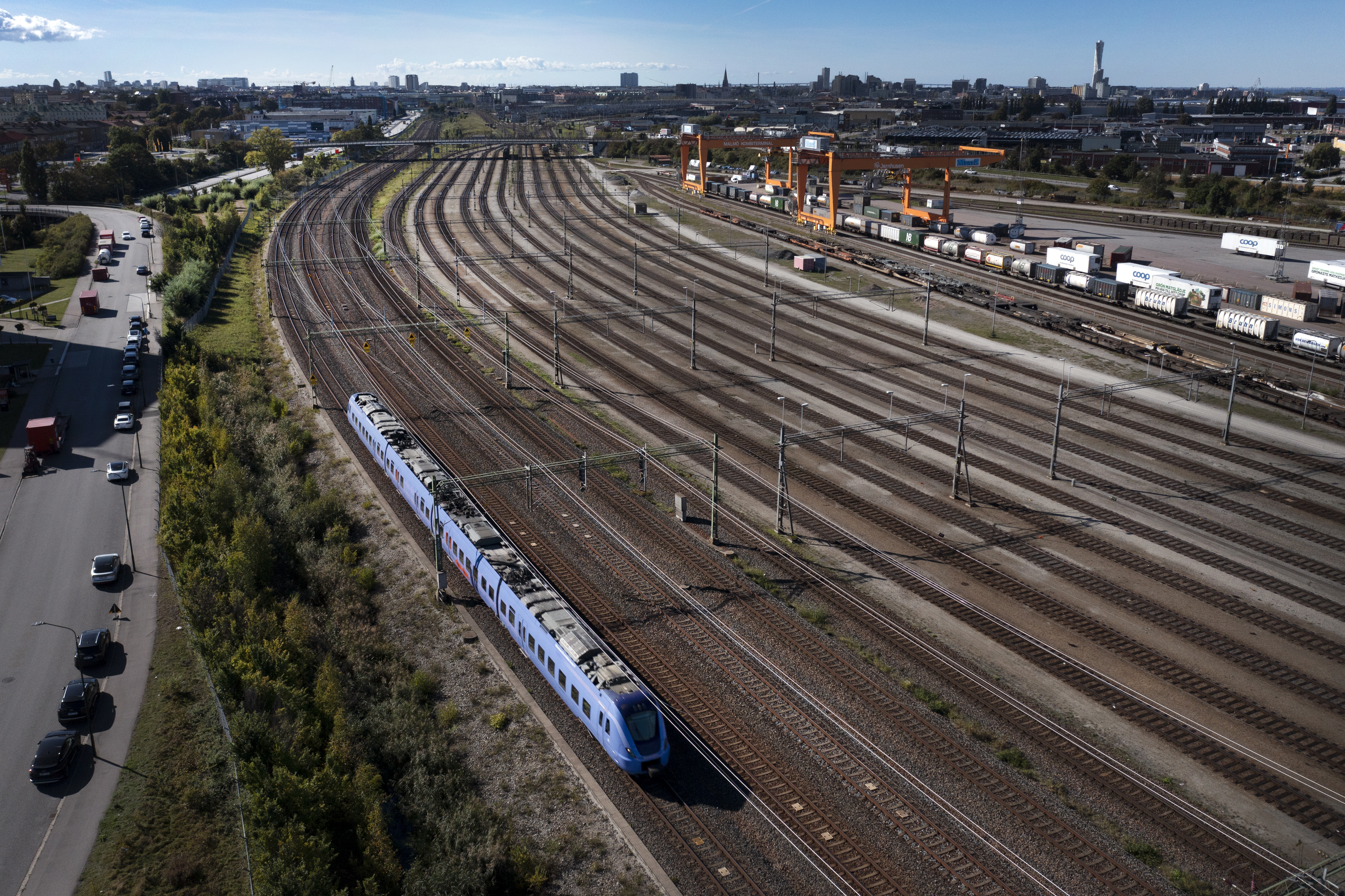 Flygvy över ett blått passagerartåg som kör på järnväg bredvid en bangård med tomma spår och containrar.